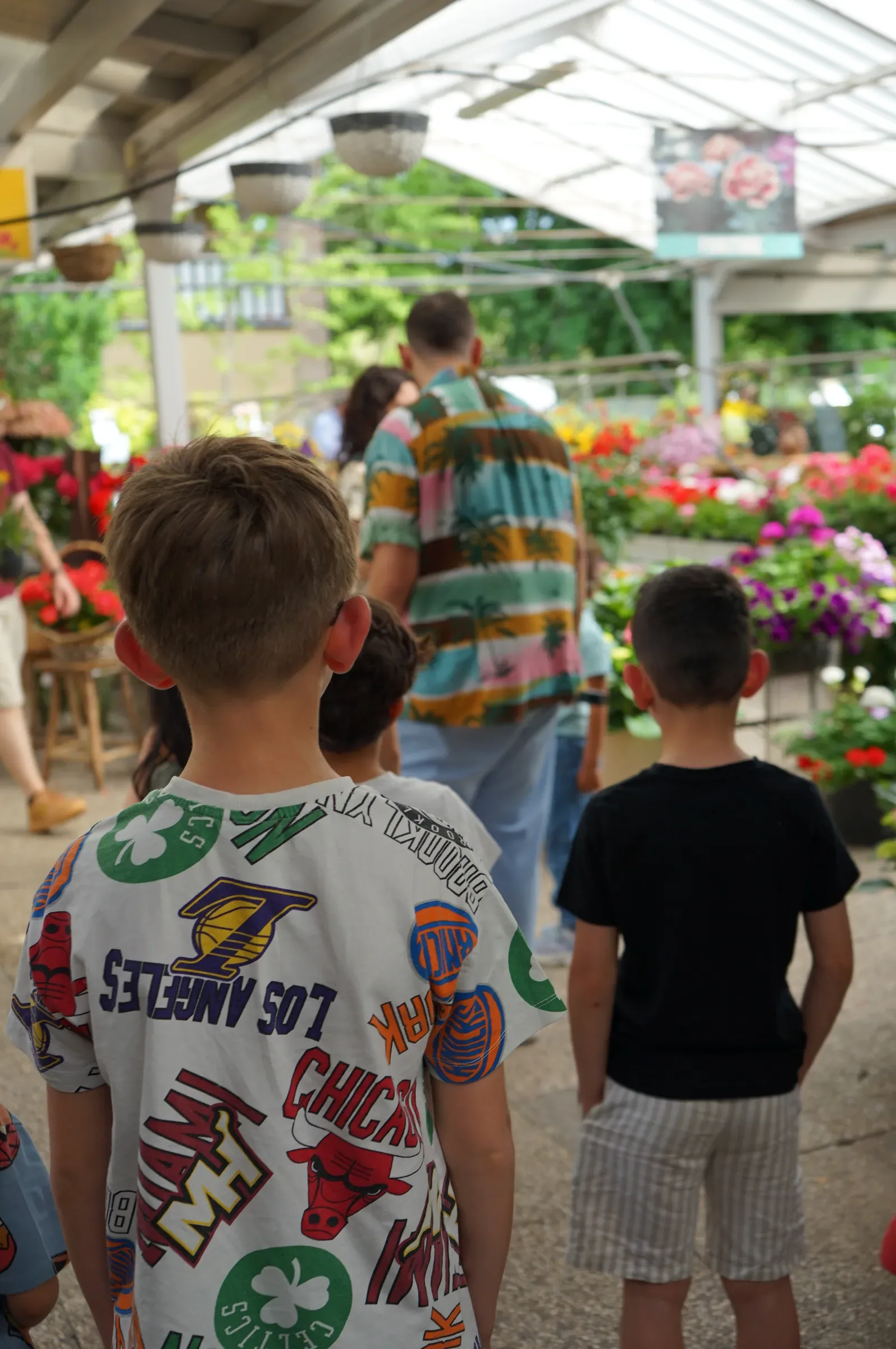 Bambini di spalle fra i banchi di fiori sotto la copertura del garden, con un adulto in camicia a fantasia
