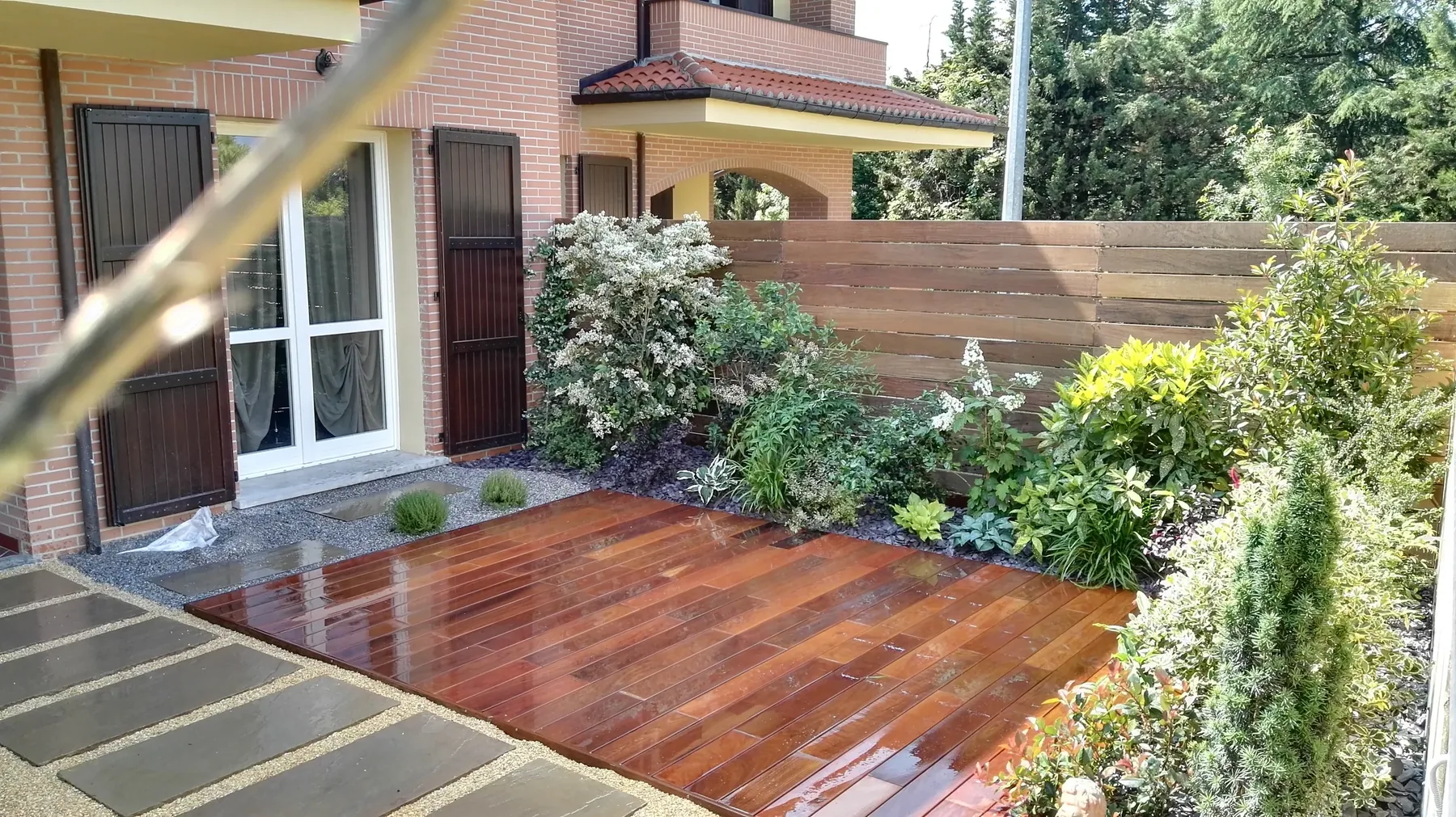 Pedana in doghe di legno bagnate e camminamento di lastre nella ghiaia, fra aiuole di arbusti davanti a una casa in mattoni