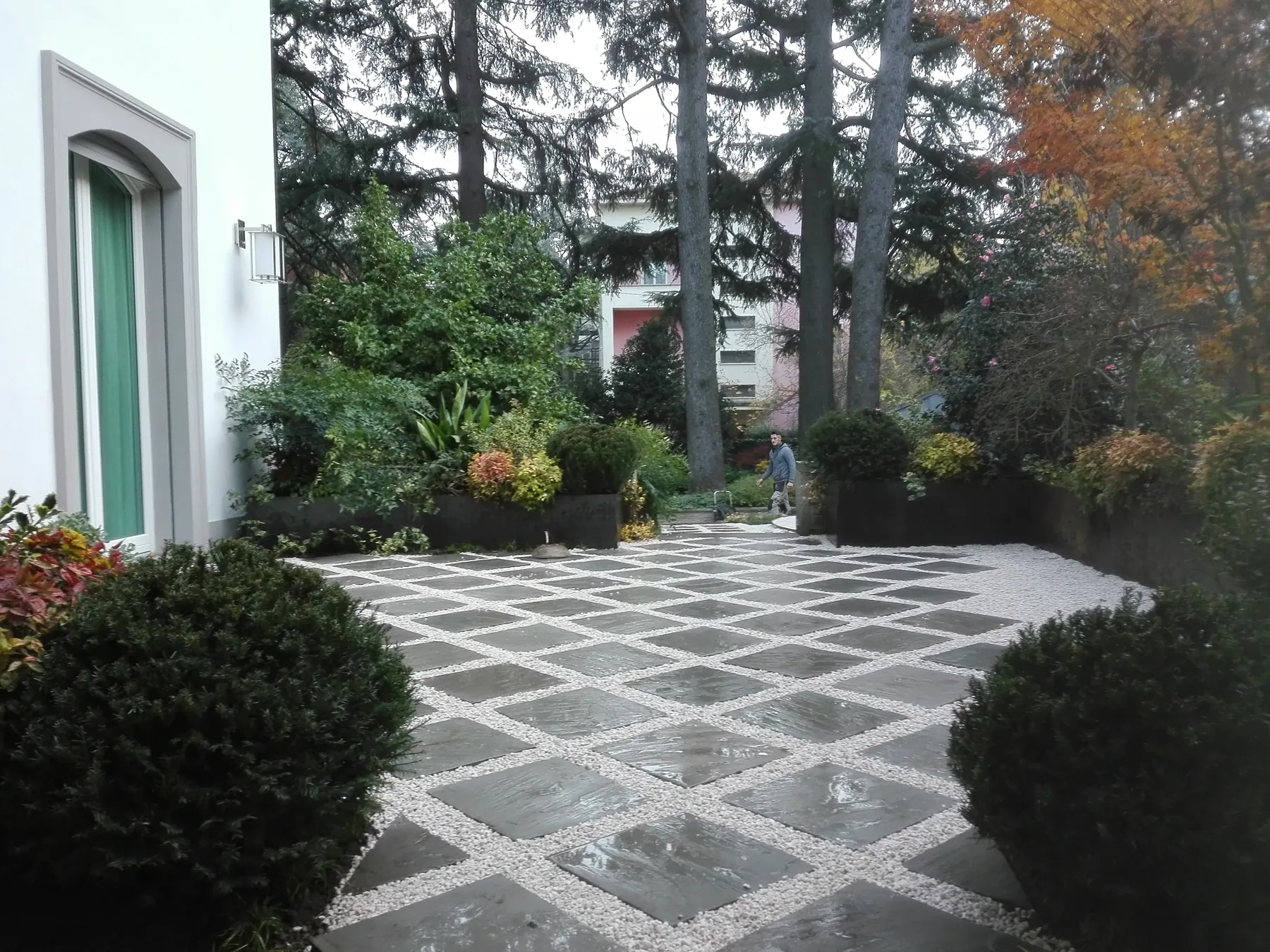 Cortile pavimentato a quadrotti di pietra scura con fughe di ghiaia bianca, fra cespugli tondi e aiuole, davanti a una casa