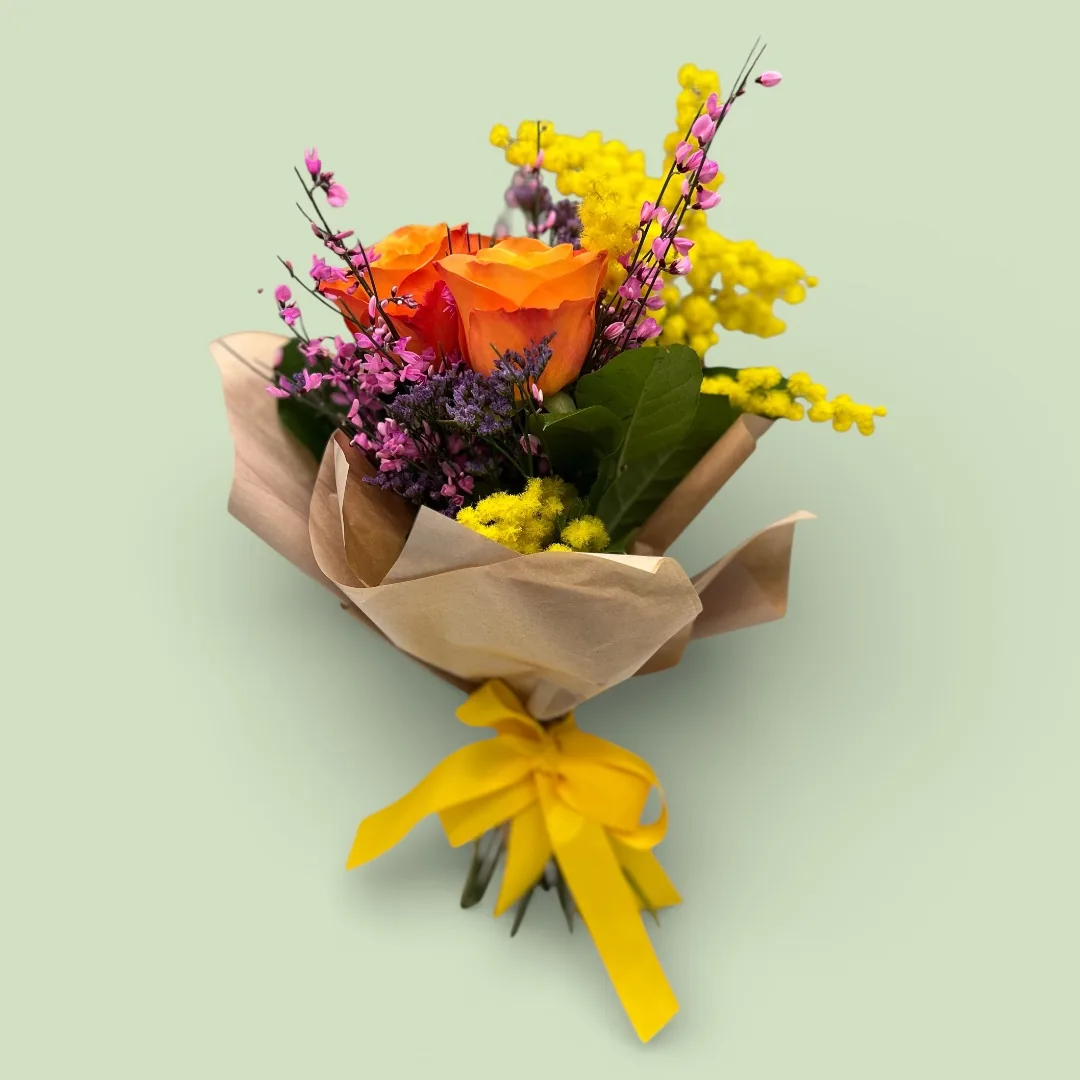 Mazzo di mimosa con rose arancio, limonium e rametti fucsia, incartato nella carta con un nastro giallo