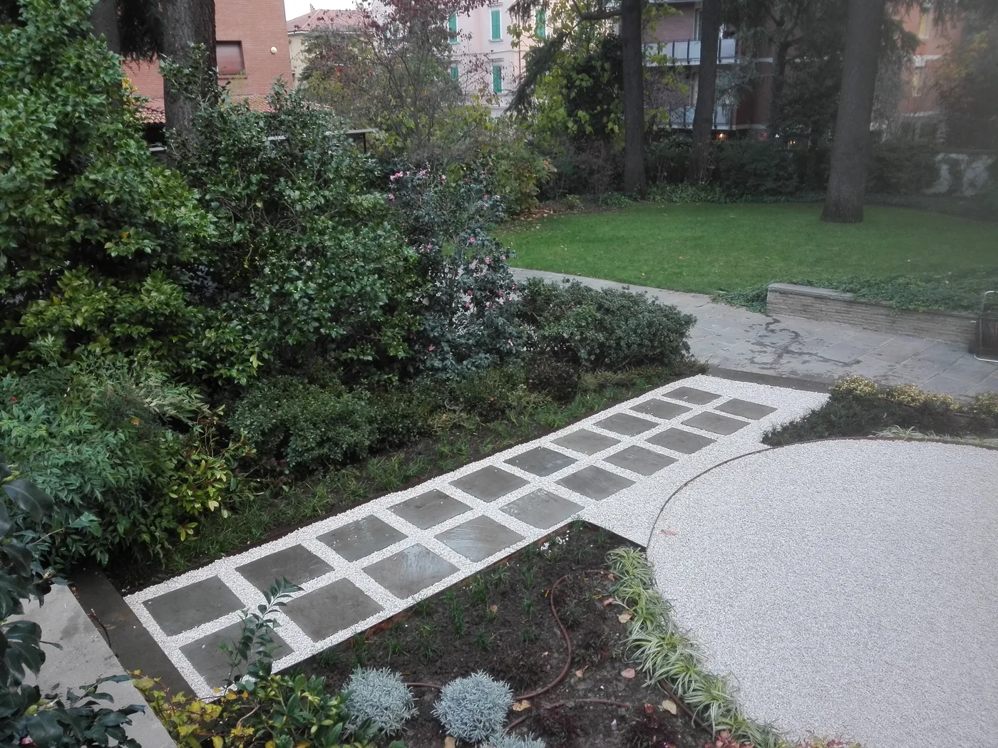 Camminamento di lastre quadrate grigio scuro bordate di graniglia bianca, che attraversa un'aiuola verso il prato, accanto a un'area tonda in ghiaia chiara