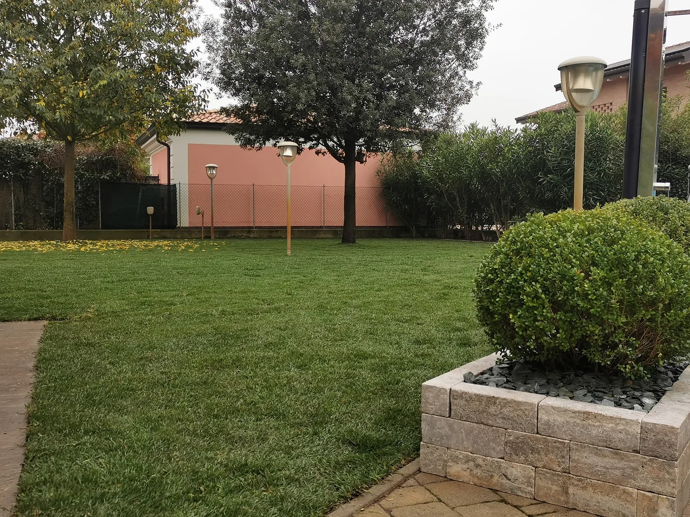 Prato davanti a una casa con un'aiuola rialzata in pietra e una sfera di bosso, e alberi sul fondo