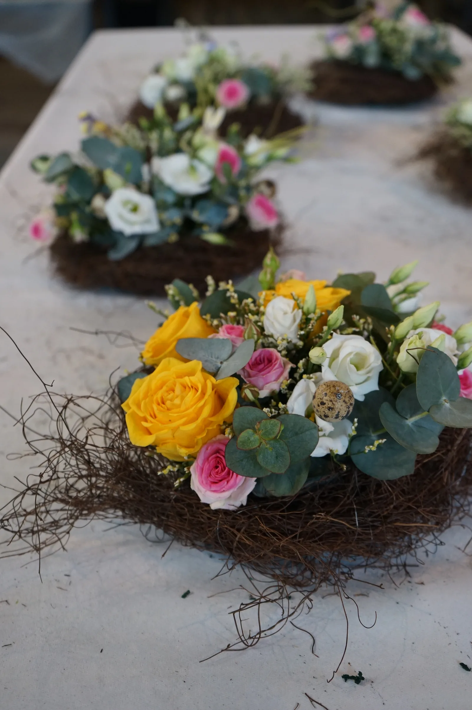 Un nido di rami con rose gialle e rosa, lisianthus bianchi ed eucalipto, e altri centrotavola sfocati sul tavolo dietro