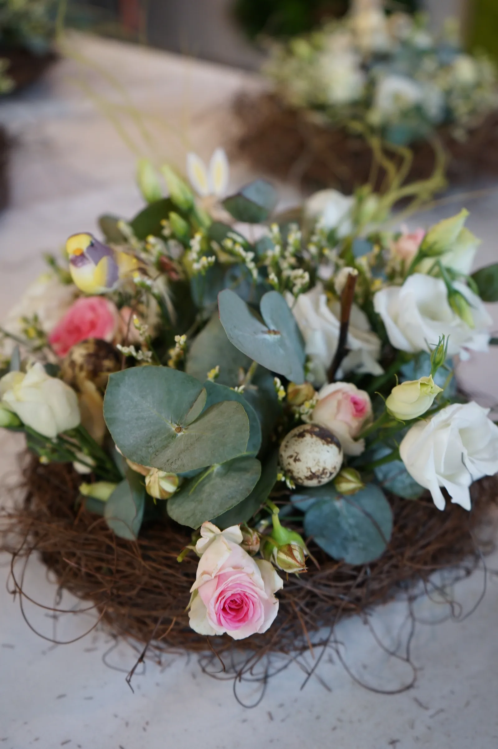 Dettaglio di un centrotavola in un nido di rametti, con foglie tonde di eucalipto, roselline rosa, lisianthus bianchi, uova di quaglia e un uccellino decorativo