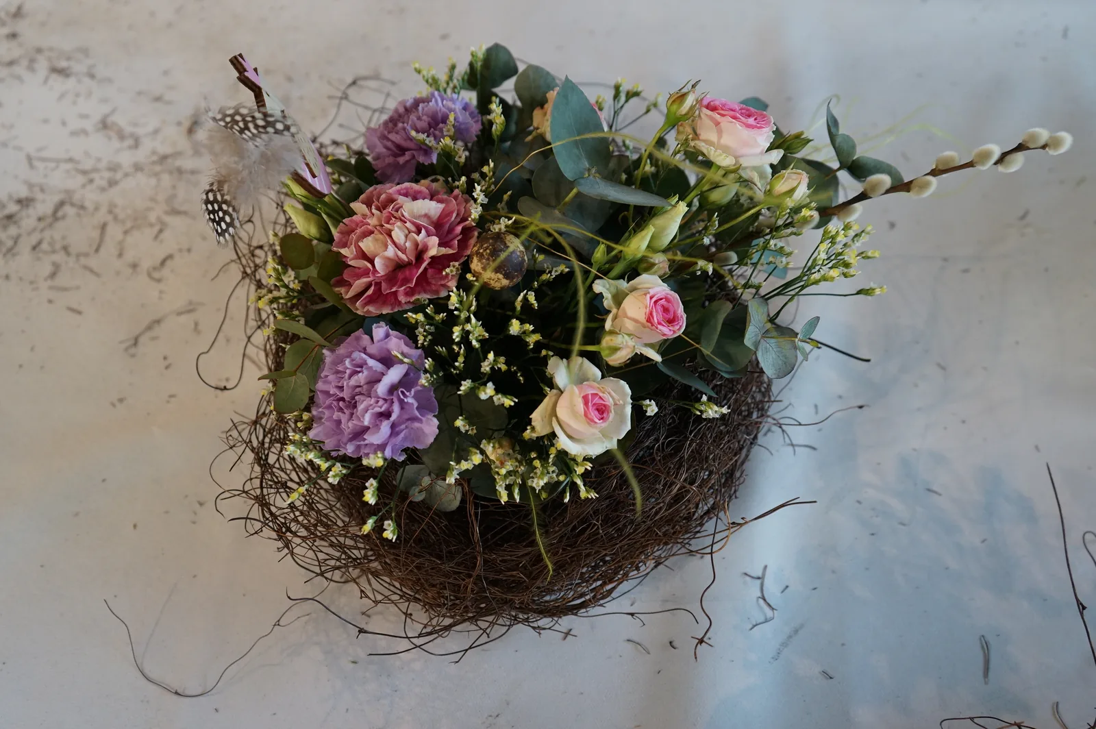 Centrotavola in un nido di rametti, visto dall'alto, con garofani viola e rosa, rose, una piuma a pois e rametti fioriti