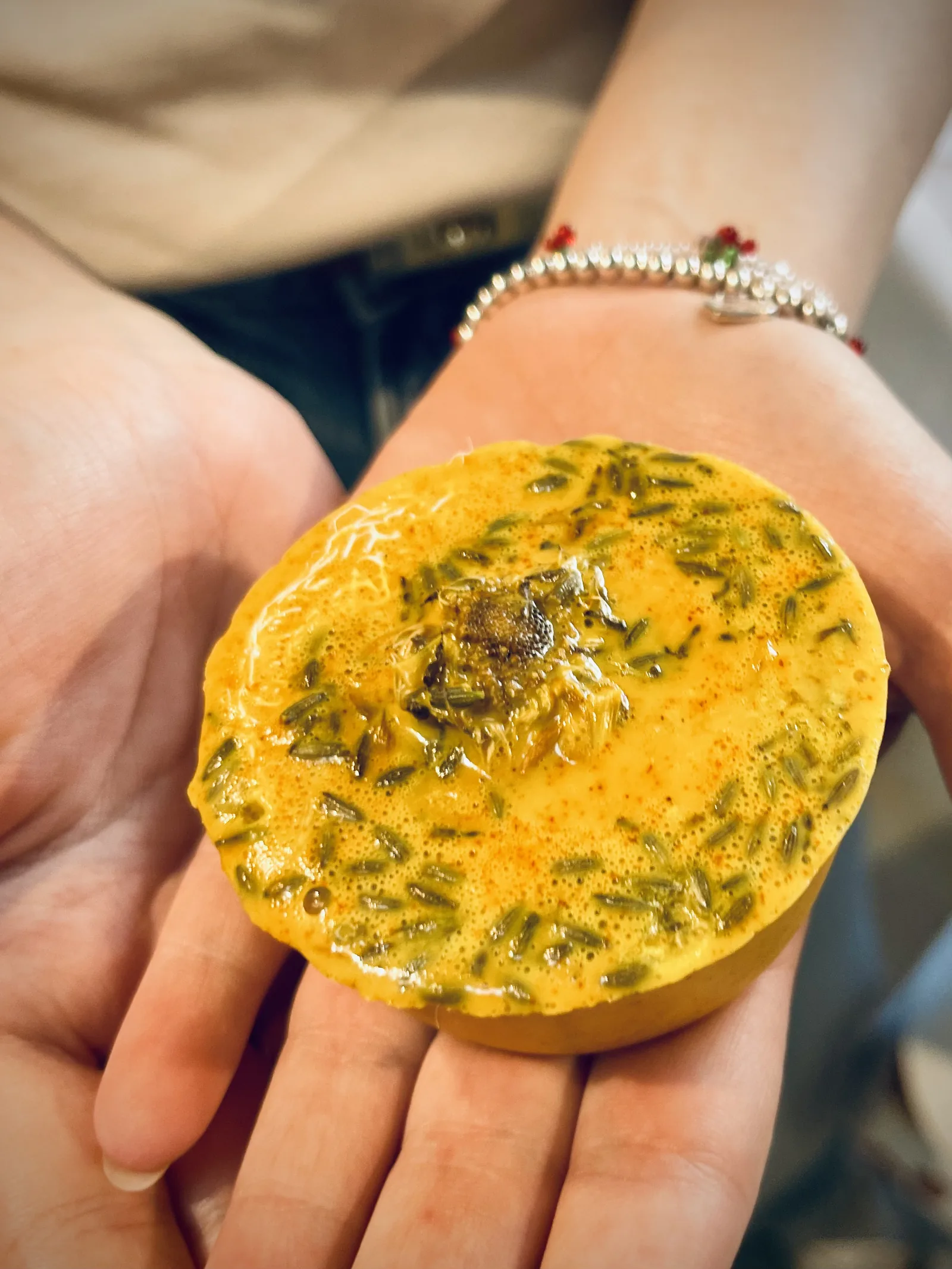 Un sapone tondo giallo arancio con semi di erbe secche e un fiore secco al centro, sul palmo di una mano