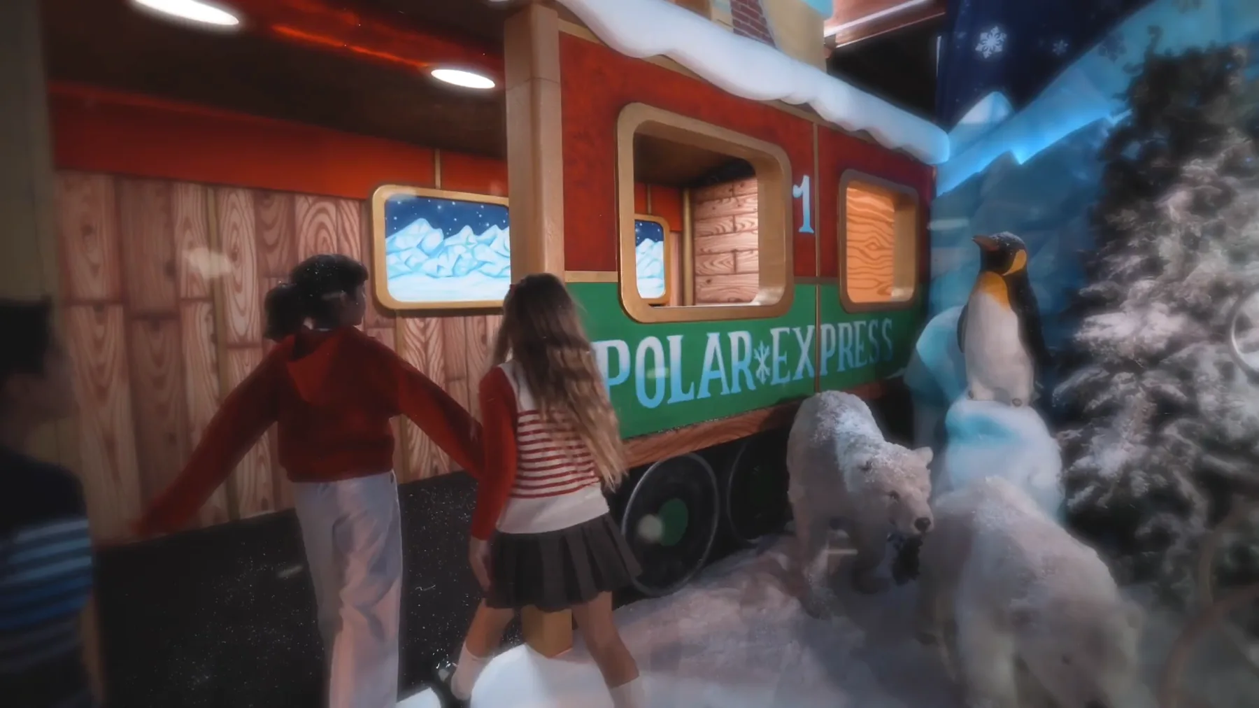 Due bambine di spalle davanti al vagone del Polar Express, fra orsi polari, un pinguino e un abete innevato