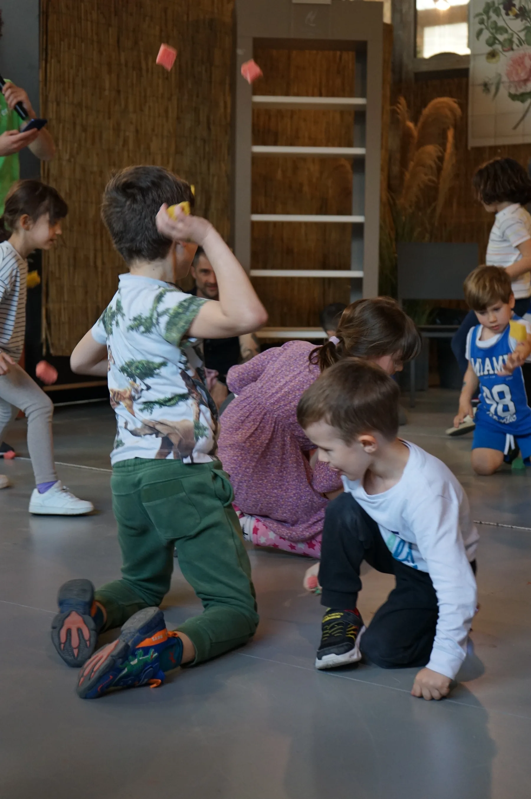 Bambini inginocchiati sul pavimento grigio; uno lancia in aria dei piccoli quadrati colorati, davanti a una parete di canne