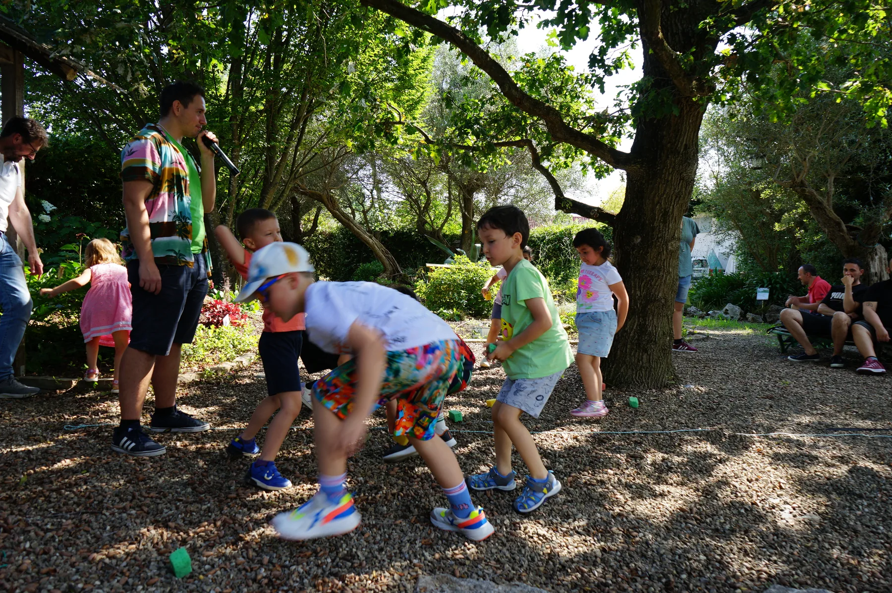 Un animatore con il microfono guida un gioco: i bambini raccolgono dalla ghiaia i cubetti di spugna verdi e gialli