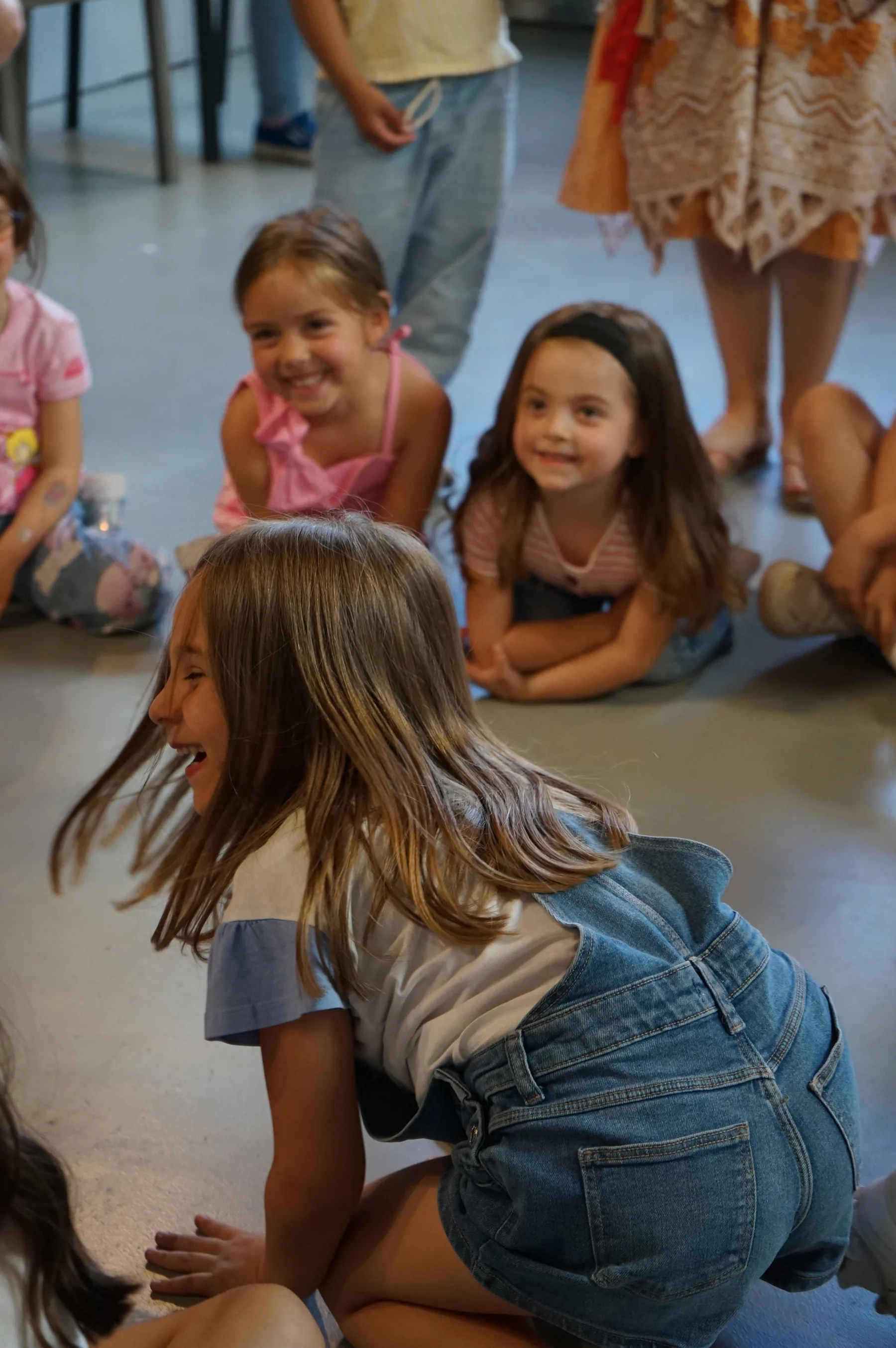 Una bambina con la salopette di jeans si gira ridendo, in ginocchio sul pavimento, davanti ad altre bambine sedute che sorridono