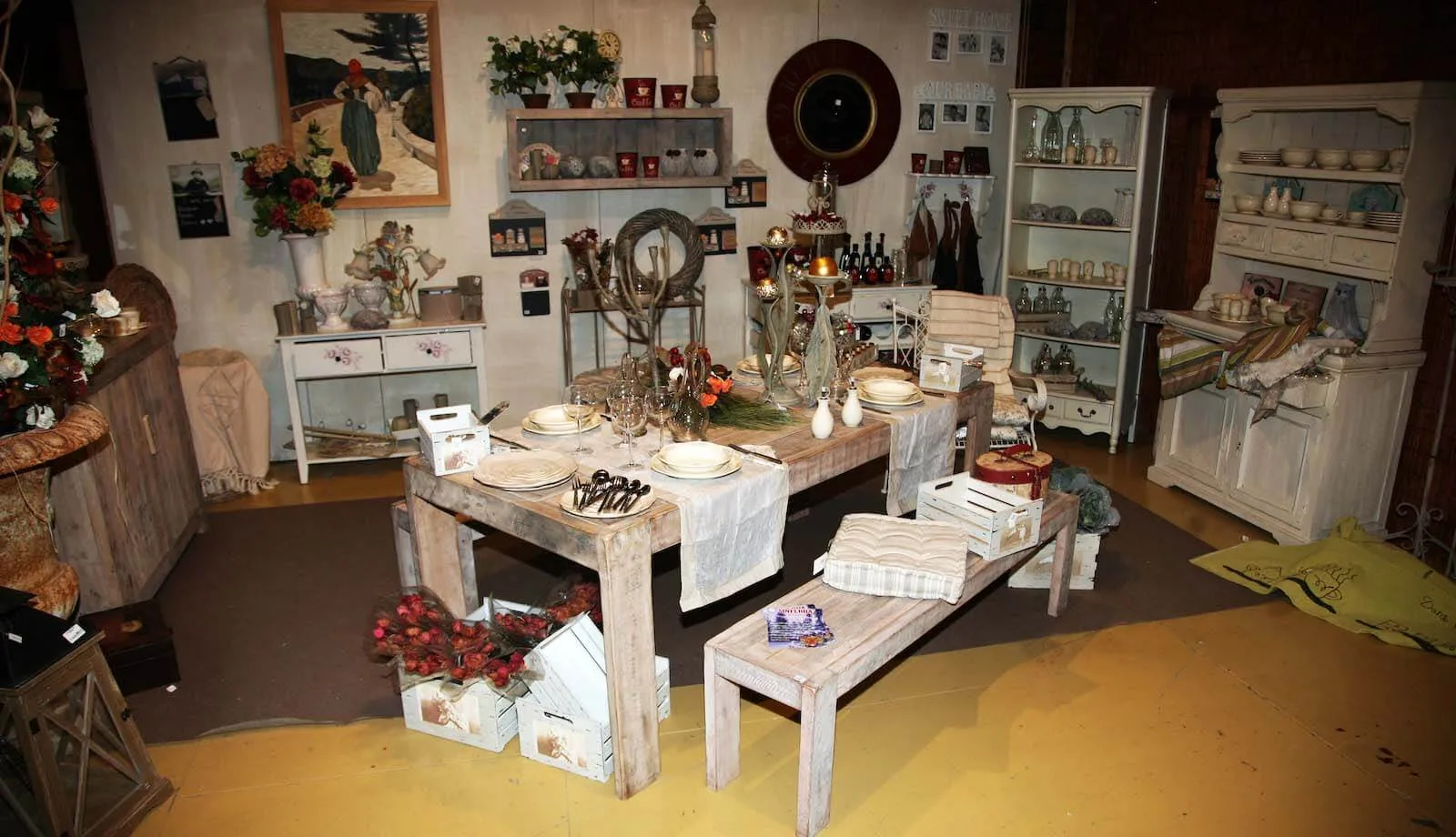Ambiente arredato in legno sbiancato, con un tavolo apparecchiato e le panche, credenze e mazzi di rose rosse nelle cassette