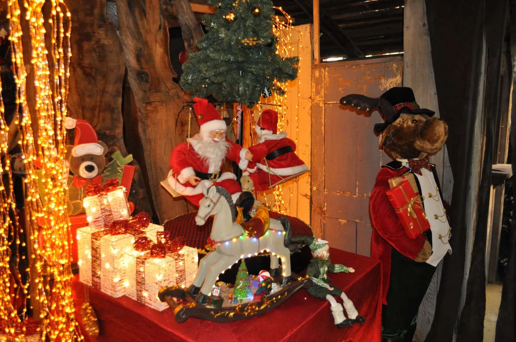 Babbo Natale e un'altra figura in rosso sulle altalene, dietro un cavallo a dondolo bianco illuminato, fra pacchi luminosi, orsetti e un folletto, accanto a un alce di peluche in giacca rossa