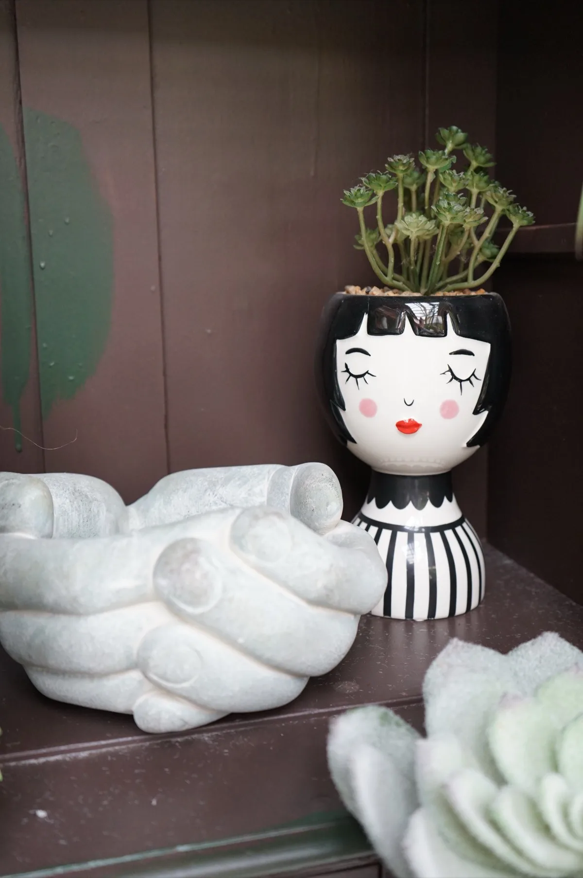 Vaso in ceramica bianca e nera a forma di volto con gli occhi chiusi e il caschetto dipinto, con una succulenta che spunta dalla testa, accanto a una ciotola a forma di mani