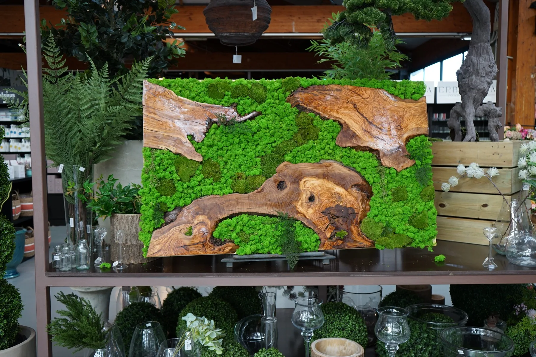 Quadro vegetale di muschio e lastre di legno venato, appoggiato su una mensola fra vetri e sfere verdi