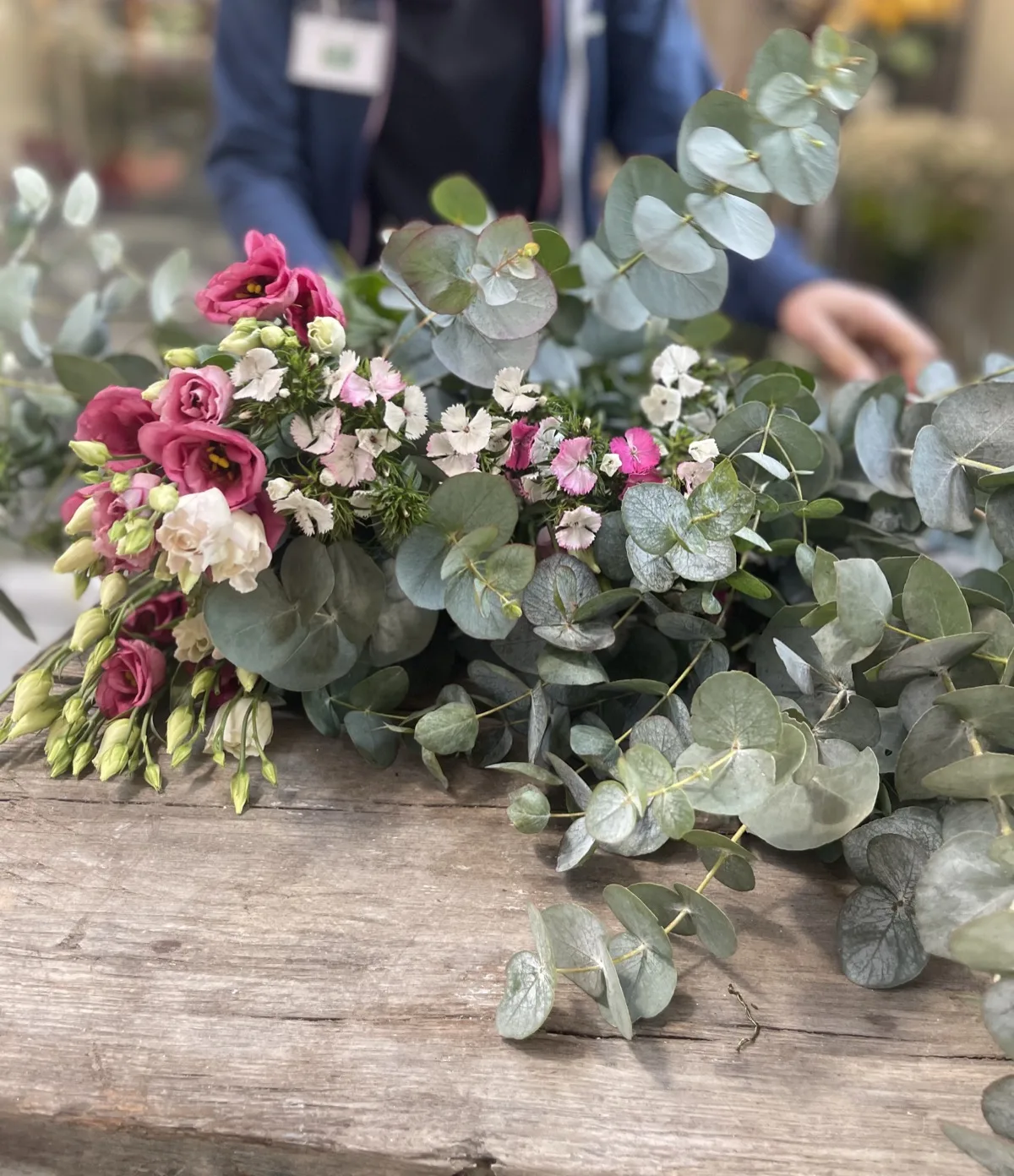 Lisianthus rosa e fiorellini bianchi e rosa appoggiati sul piano di legno del banco, in mezzo a rami di eucalipto, con la fiorista sullo sfondo