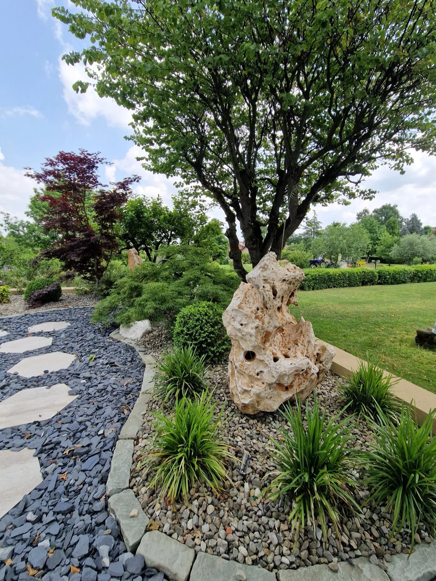 Roccia ornamentale chiara e traforata in un'aiuola di ciottoli con ciuffi di graminacee, accanto a un sentiero di scaglie scure e passi in pietra, sotto un albero