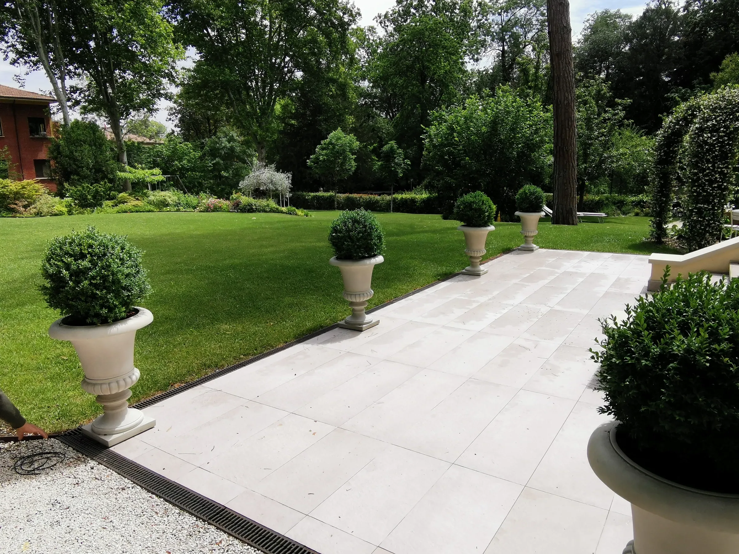 Giardino di una villa con il prato all'inglese, una terrazza in lastre di pietra chiara e sfere di bosso in vasi a coppa, fra gli alberi alti del parco