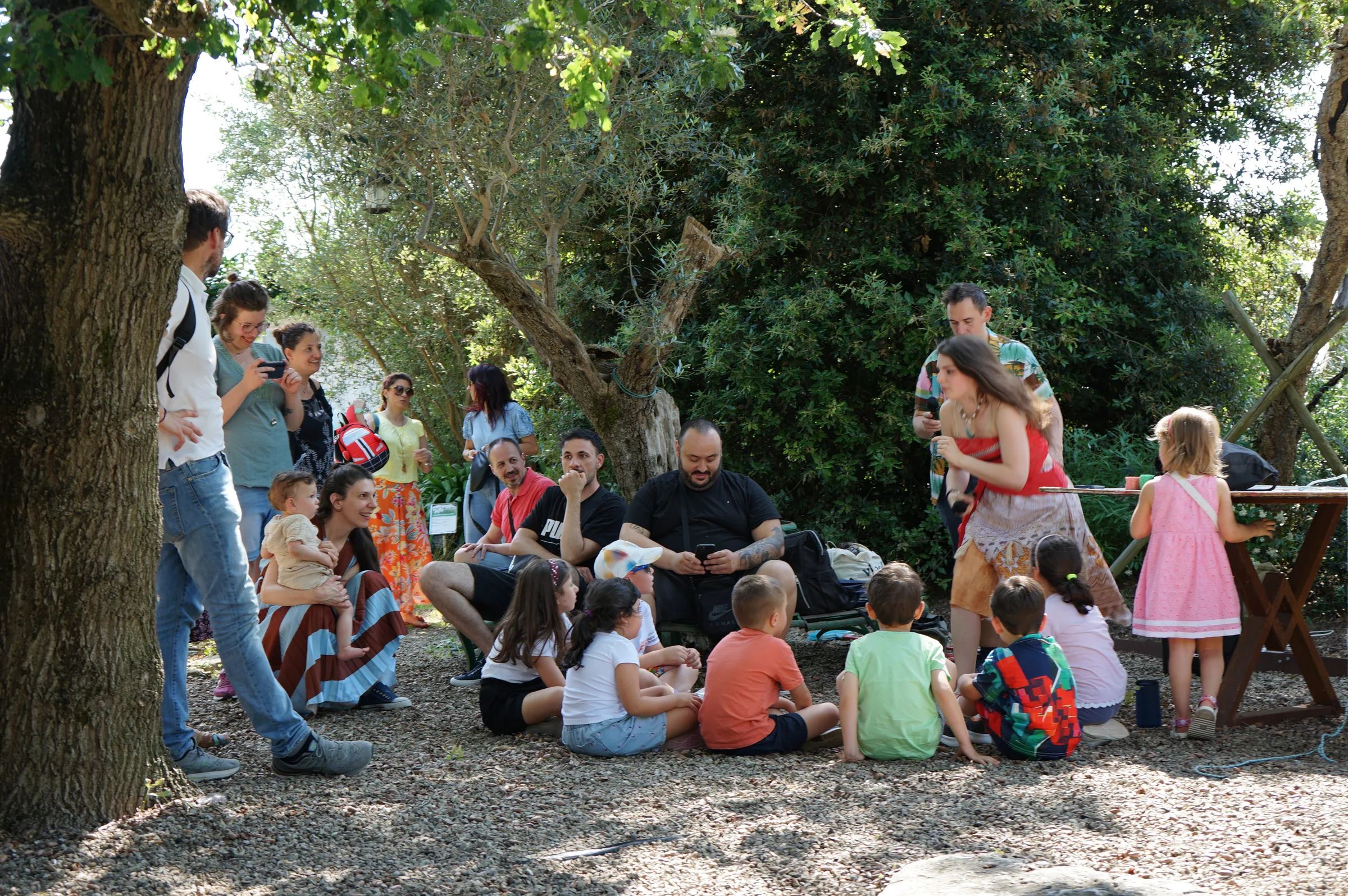 Bambini seduti sulla ghiaia sotto gli alberi, con una ragazza in costume e gli adulti intorno