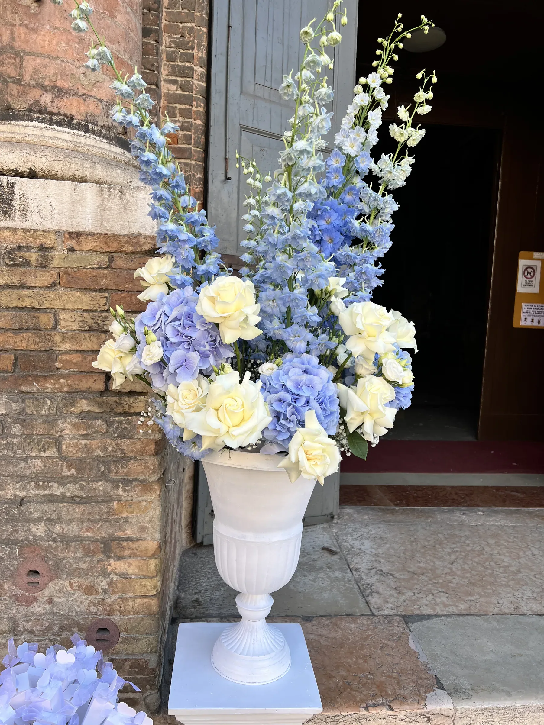 Composizione alta di delphinium azzurri, ortensie azzurre e rose bianche in un vaso a calice bianco su un piedistallo, sui gradini di pietra accanto a un muro di mattoni e a un portone aperto