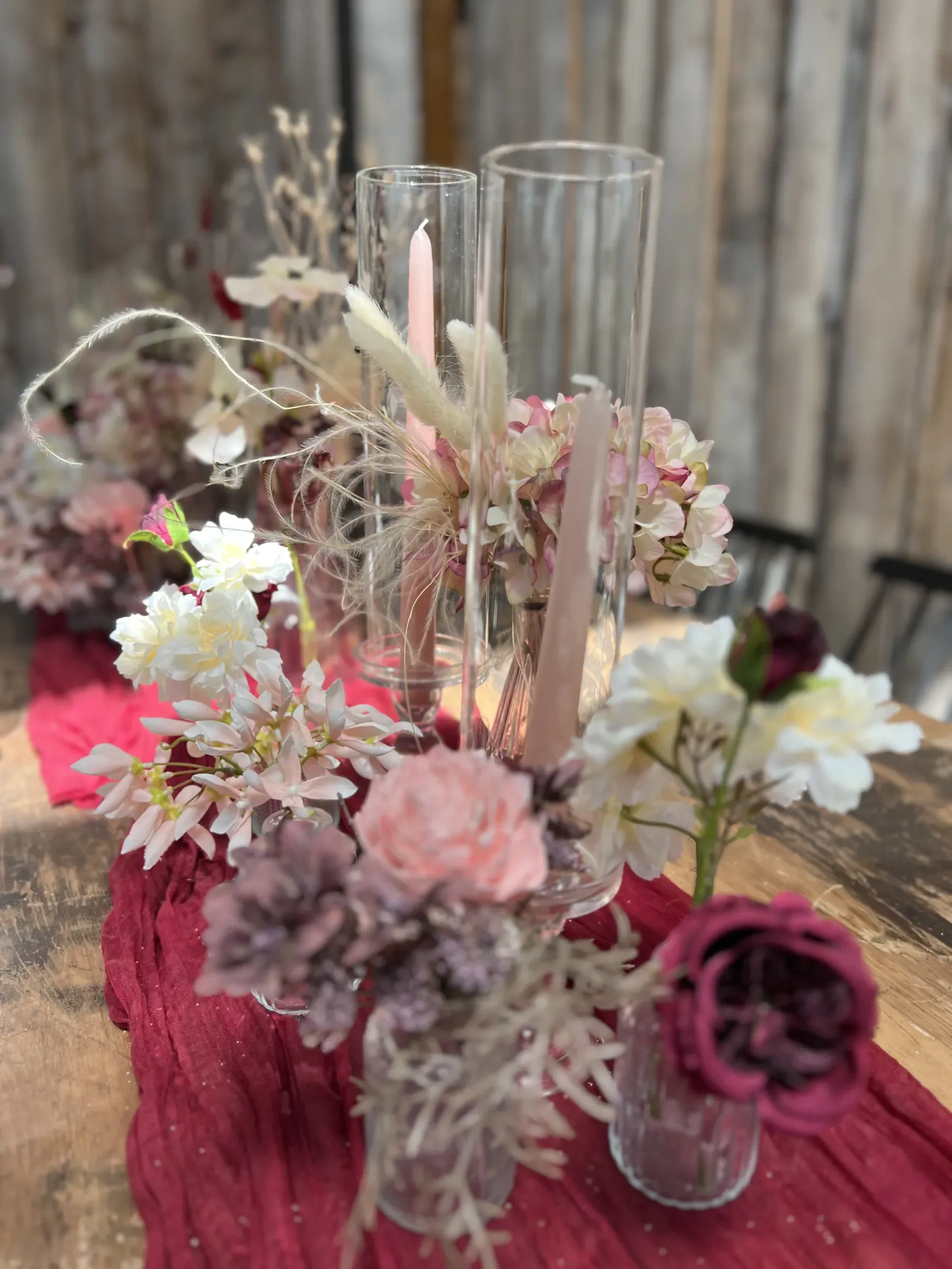Centrotavola in vasetti di vetro con ortensie rosa, fiori bianchi, una rosa cipria, una bordeaux e spighe secche, con candele rosa e cilindri di vetro su un runner bordeaux