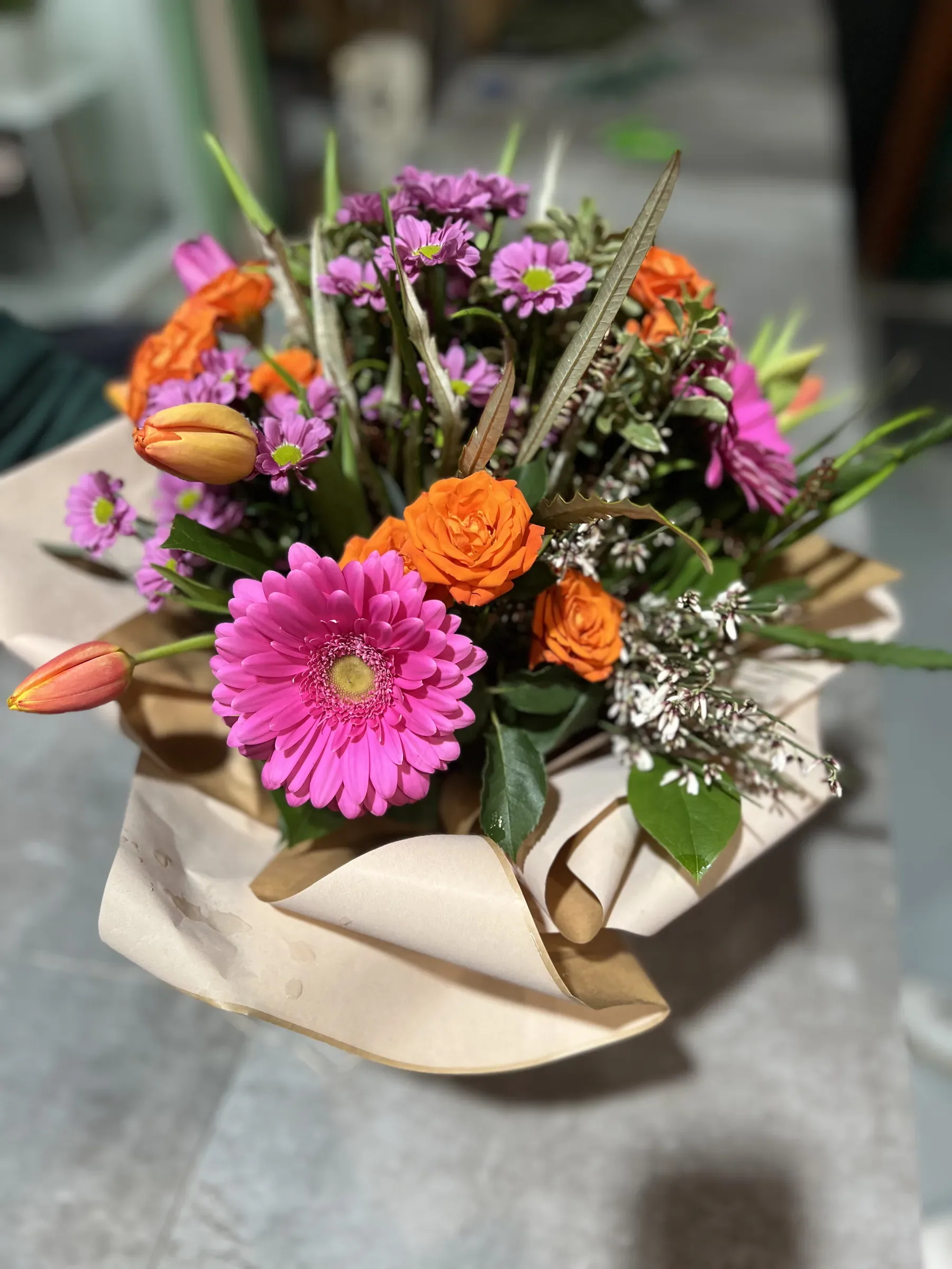 Composizione di gerbera fucsia, rose arancio, tulipani e fiori lilla in un involucro di carta color sabbia