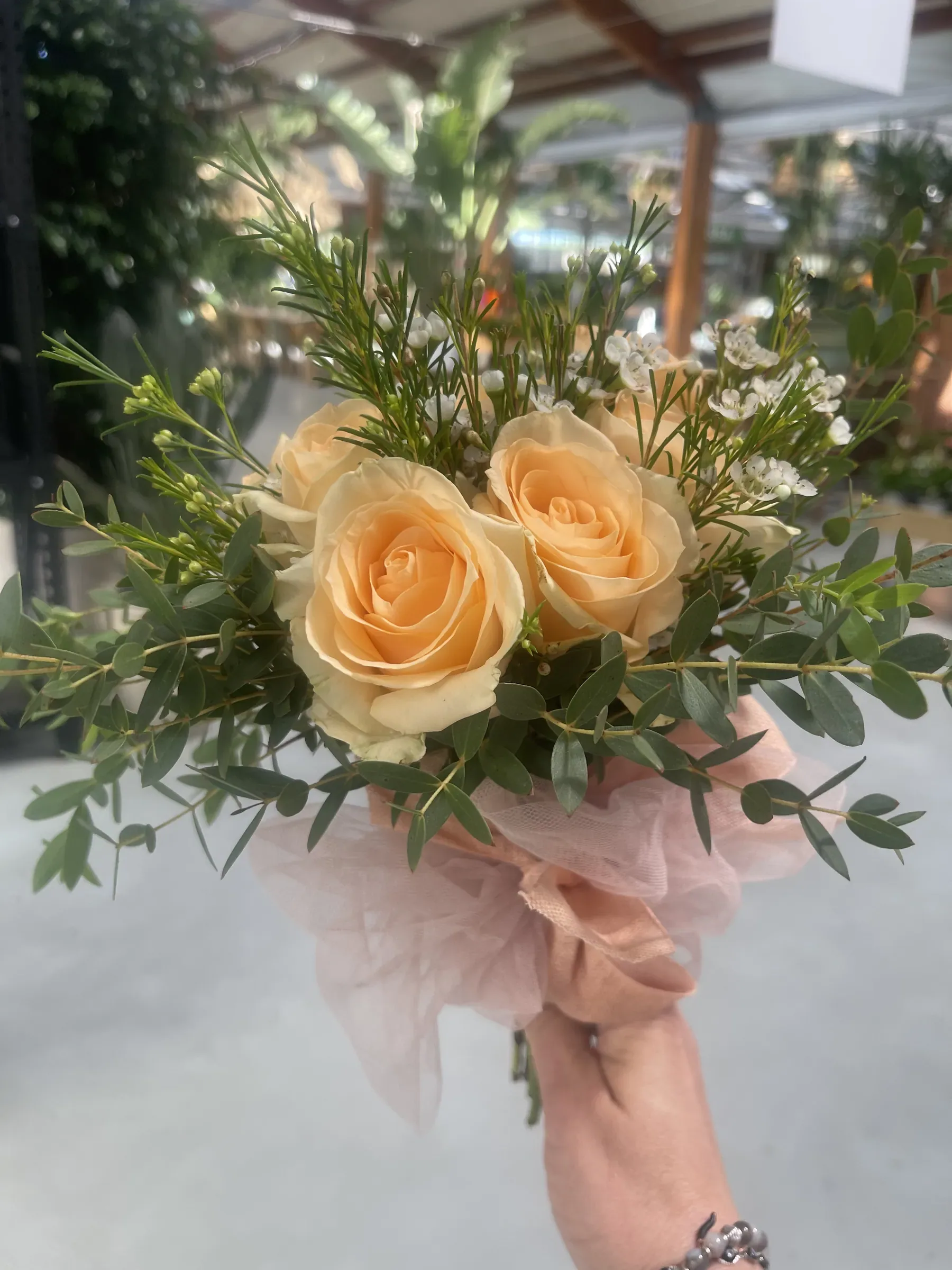 Mazzolino di rose color pesca con rametti verdi, legato con un tulle rosa e tenuto in mano sotto la serra