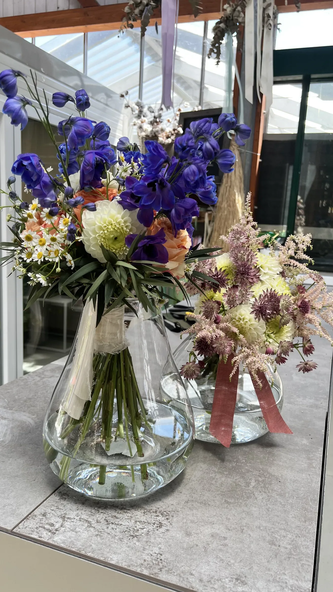 Due vasi di vetro con delphinium blu, fiori bianchi e rosa pallido e nastri, su un piano grigio in serra