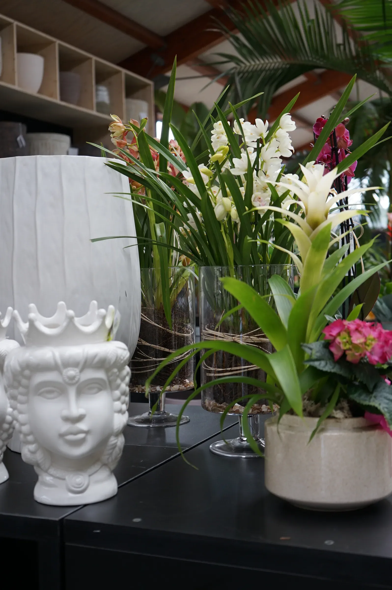 Vasi bianchi a forma di testa con la corona, accanto a un grande vaso bianco, orchidee in vasi di vetro e una bromelia bianca con un'ortensia rosa