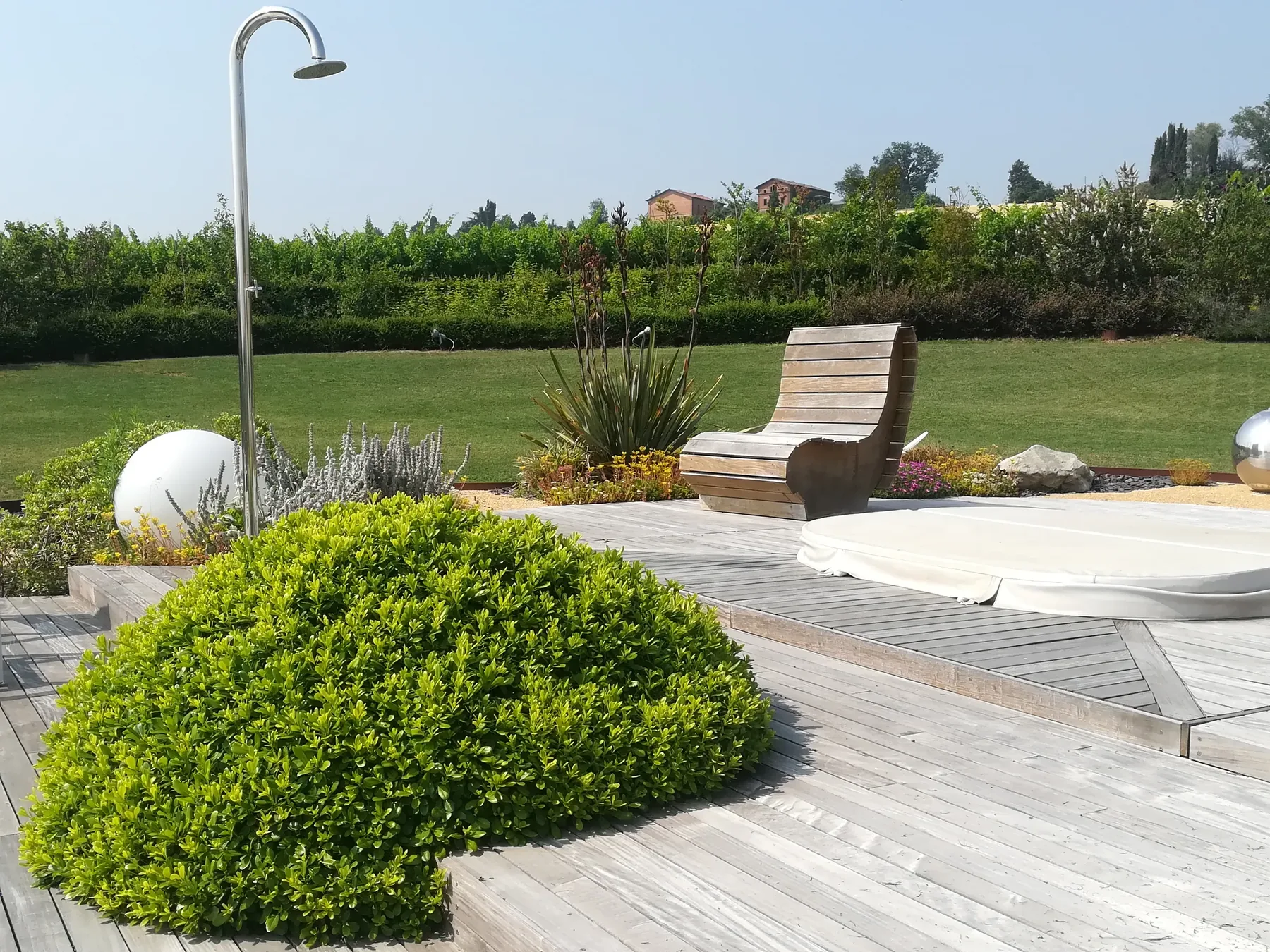 Pedana in legno attorno a un cespuglio tondo, con una sfera bianca, una doccia da giardino e una seduta in legno