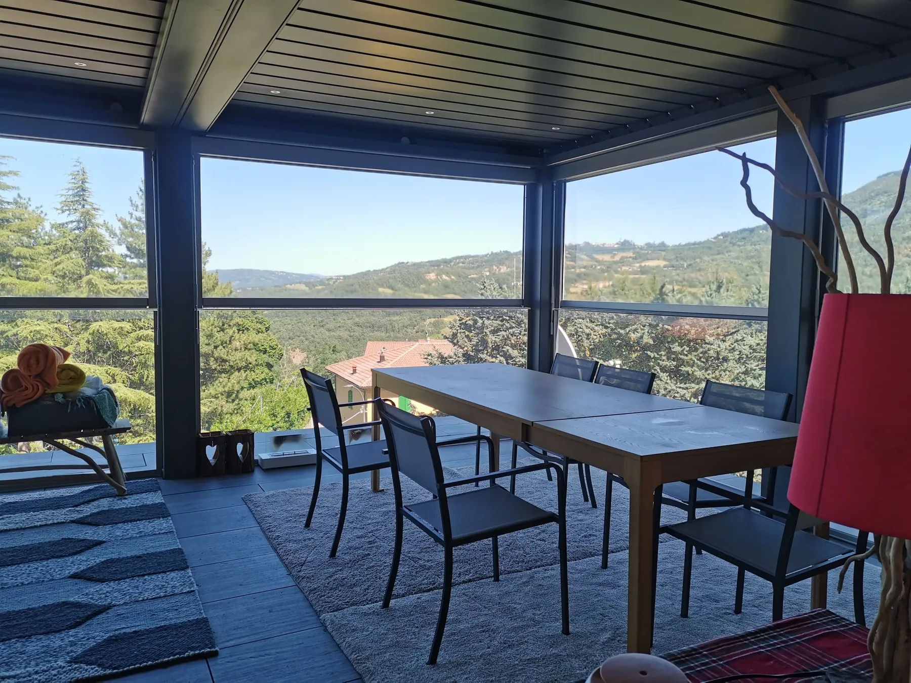 Pergola con la copertura a lamelle chiusa e le pareti vetrate affacciate sulle colline, con un tavolo lungo su un tappeto chiaro
