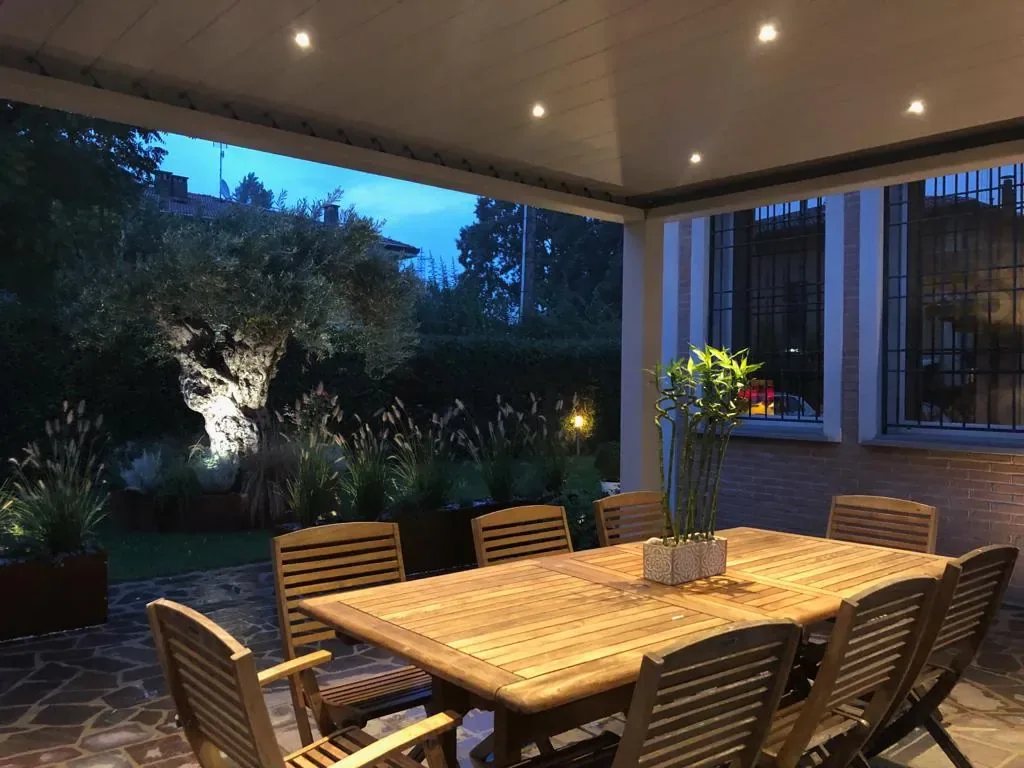 Pergola di sera con i faretti accesi nel soffitto, un tavolo in legno con le sedie e un olivo illuminato in giardino