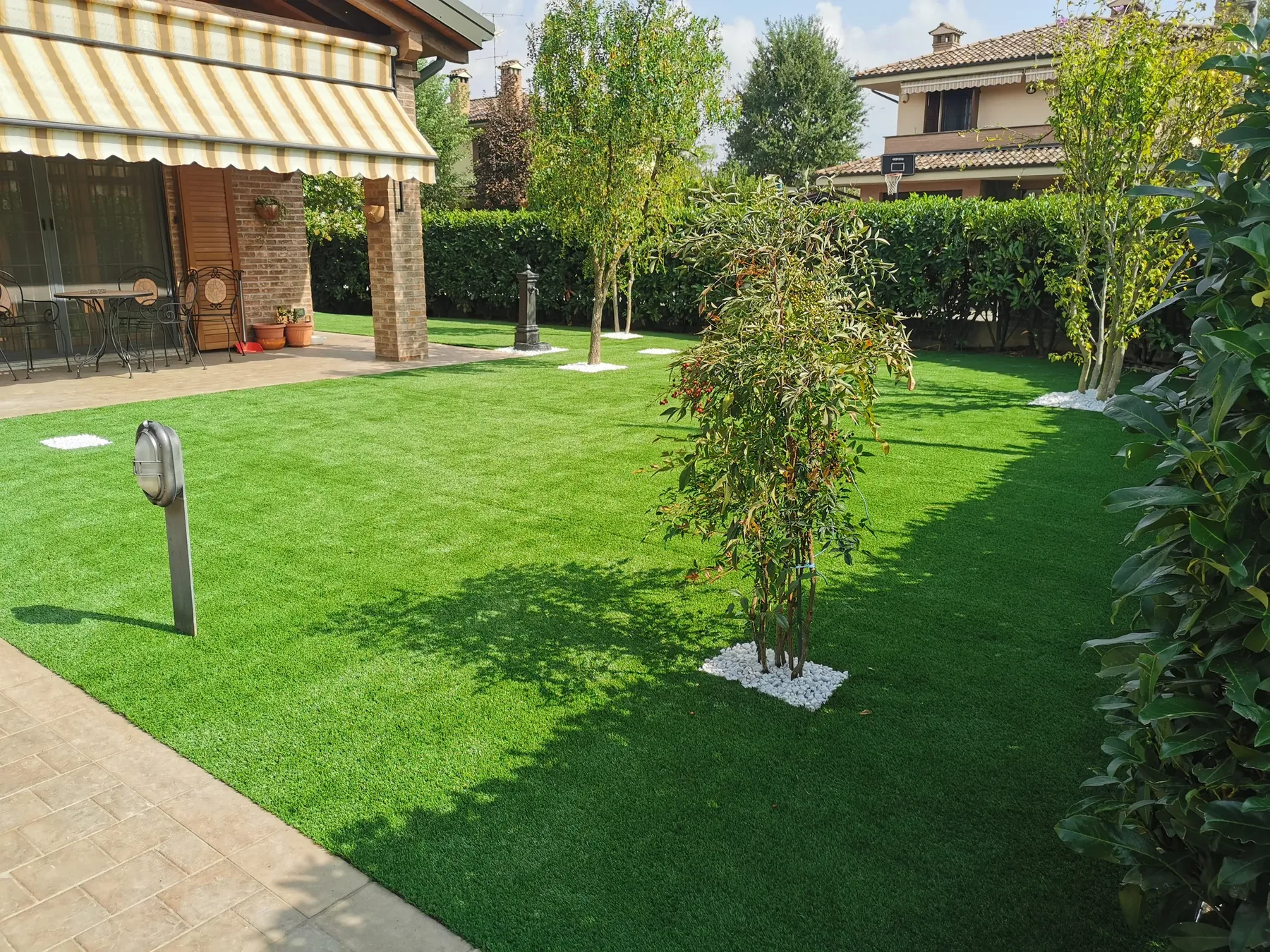 Prato sintetico ampio con alberelli giovani su quadrati di ghiaia bianca, un lampione da giardino e un portico con la tenda a righe
