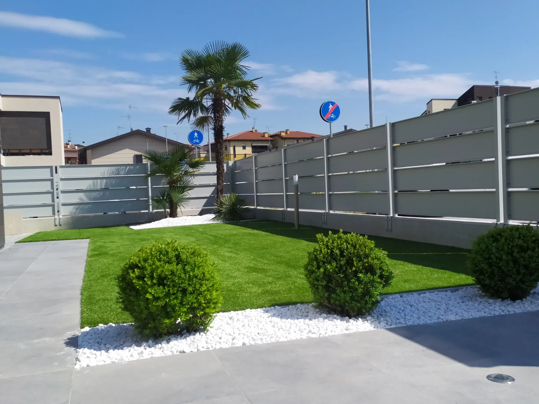 Prato sintetico con due palme su ciottoli bianchi e sfere di bosso, chiuso da una recinzione grigia a doghe