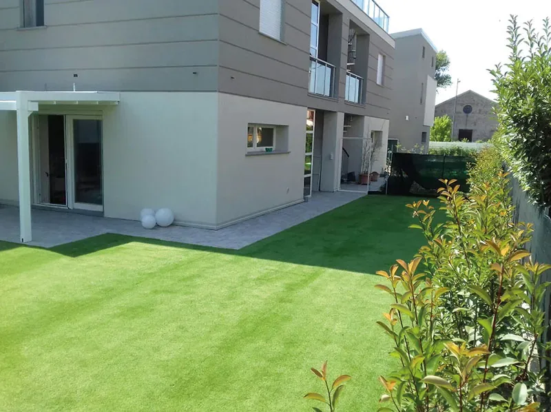 Prato sintetico davanti a una palazzina moderna con una pergola bianca, bordato da arbusti dalle foglie rosse