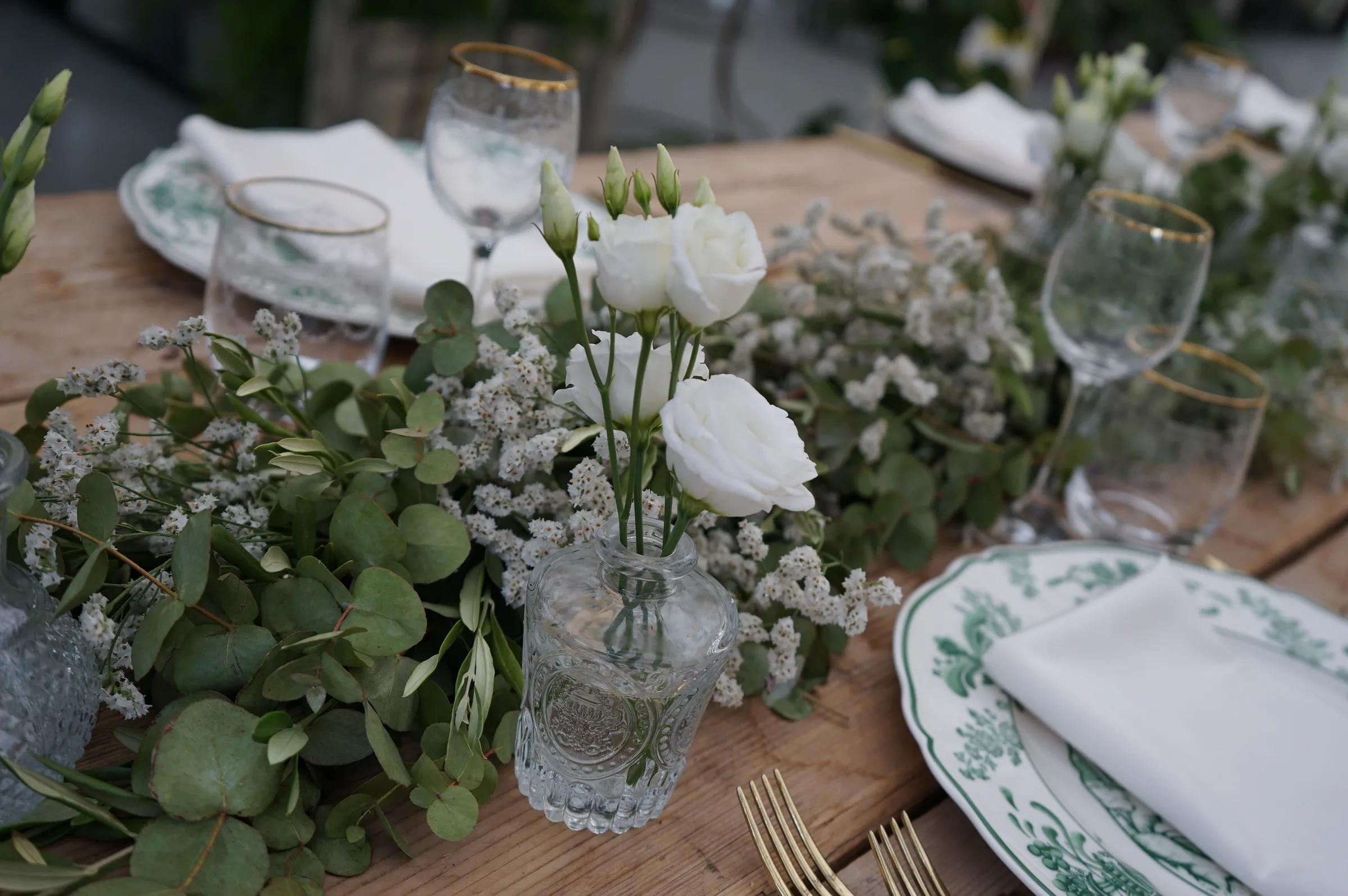 Lisianthus bianchi in un vasetto di vetro lungo il tavolo, fra rami di eucalipto e limonium bianco, accanto al piatto a decoro verde
