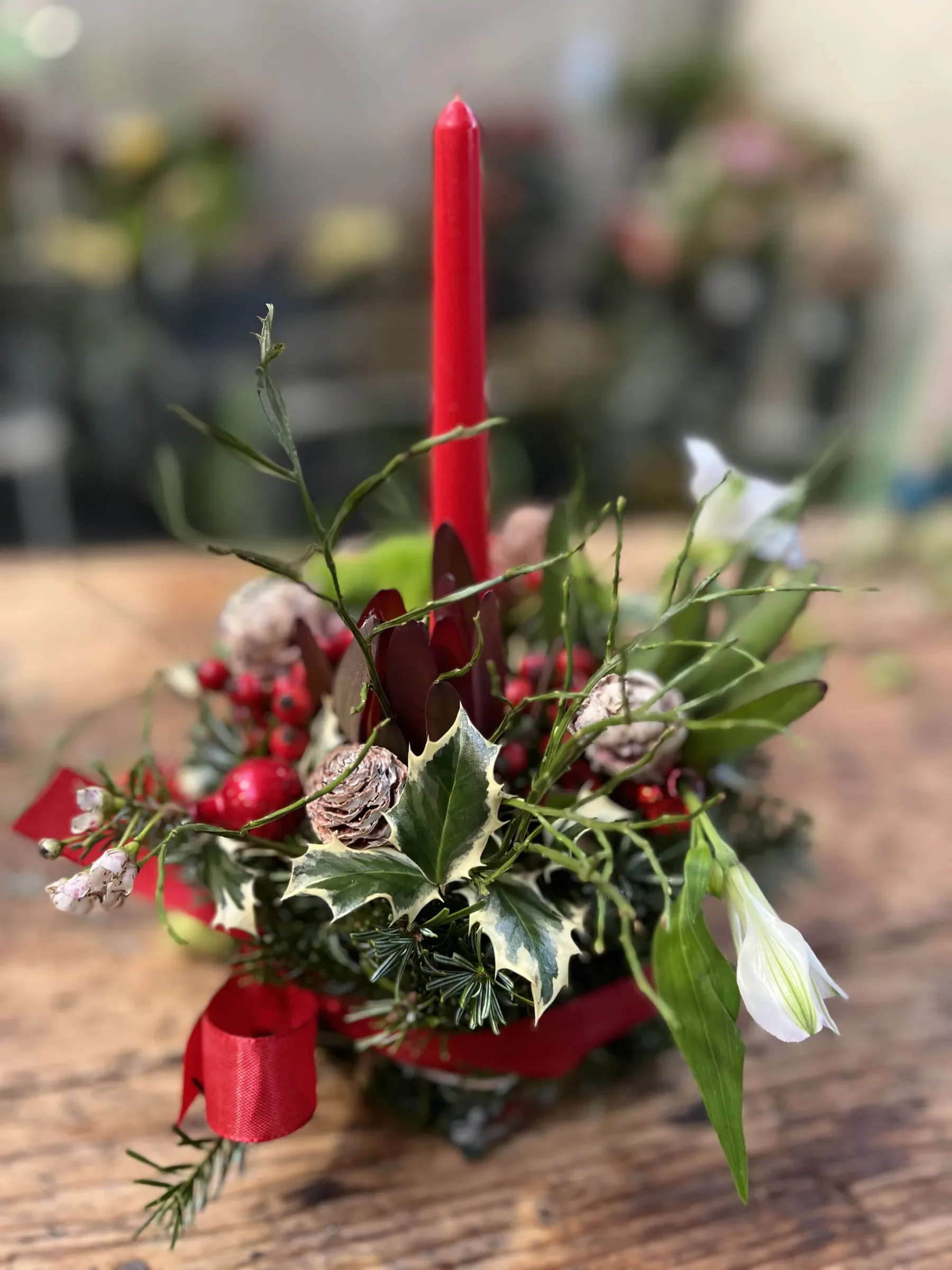 Centrotavola di Natale con una candela rossa, agrifoglio variegato, pigne, bacche rosse e un nastro rosso, sul tavolo di legno