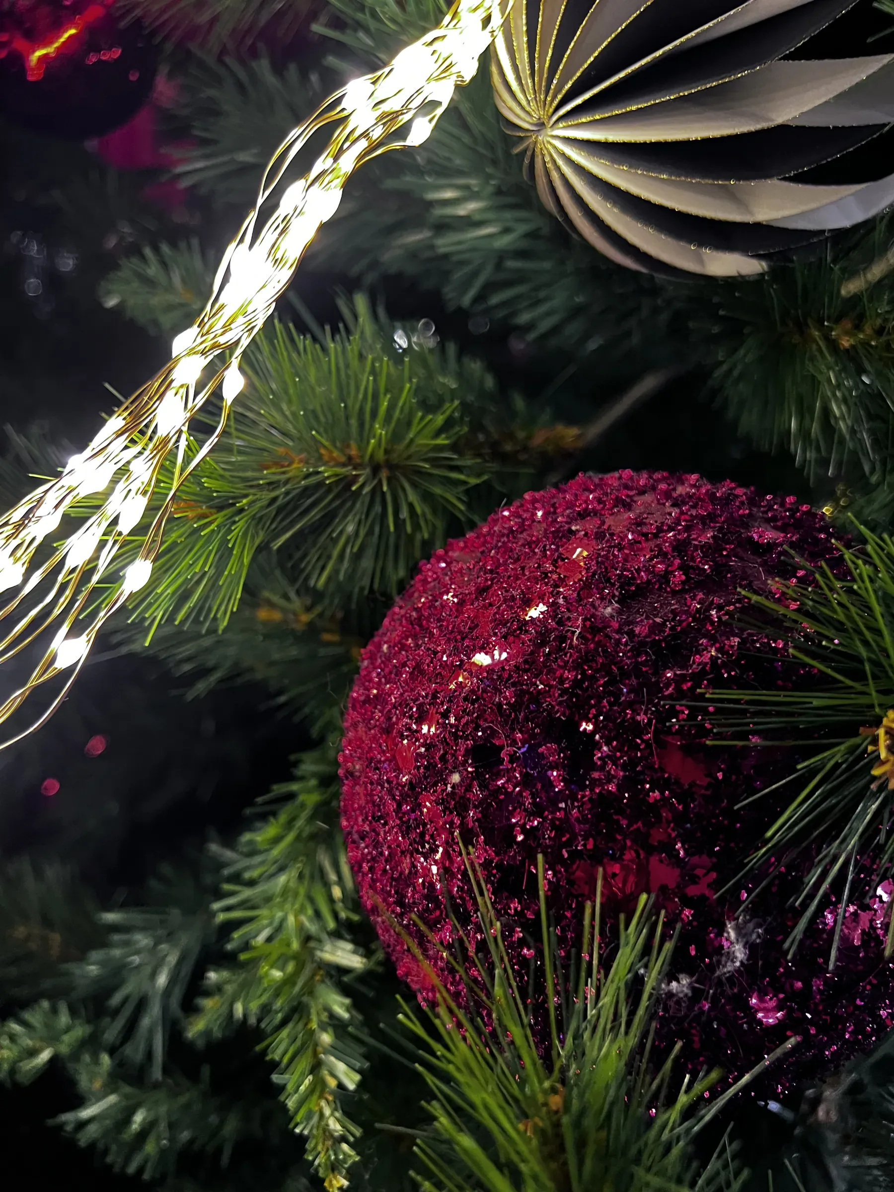 Pallina bordeaux coperta di glitter fra i rami di un abete, con un filo di lucine