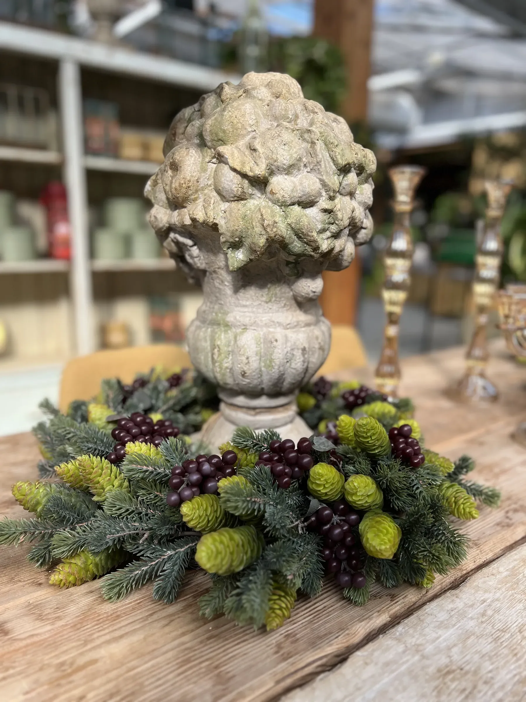 Scultura color pietra a forma di coppa colma di frutti e foglie, al centro di una corona di rametti d'abete, bacche scure e luppolo, su un tavolo di legno