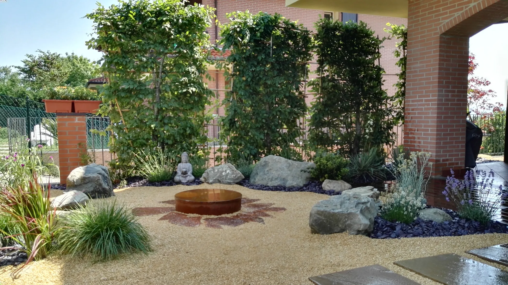 Cortile d'ingresso con pannelli di piante allevate a spalliera su grigliati di canne, una vasca tonda in corten sulla ghiaia, una statuina di Buddha, massi e graminacee ai lati