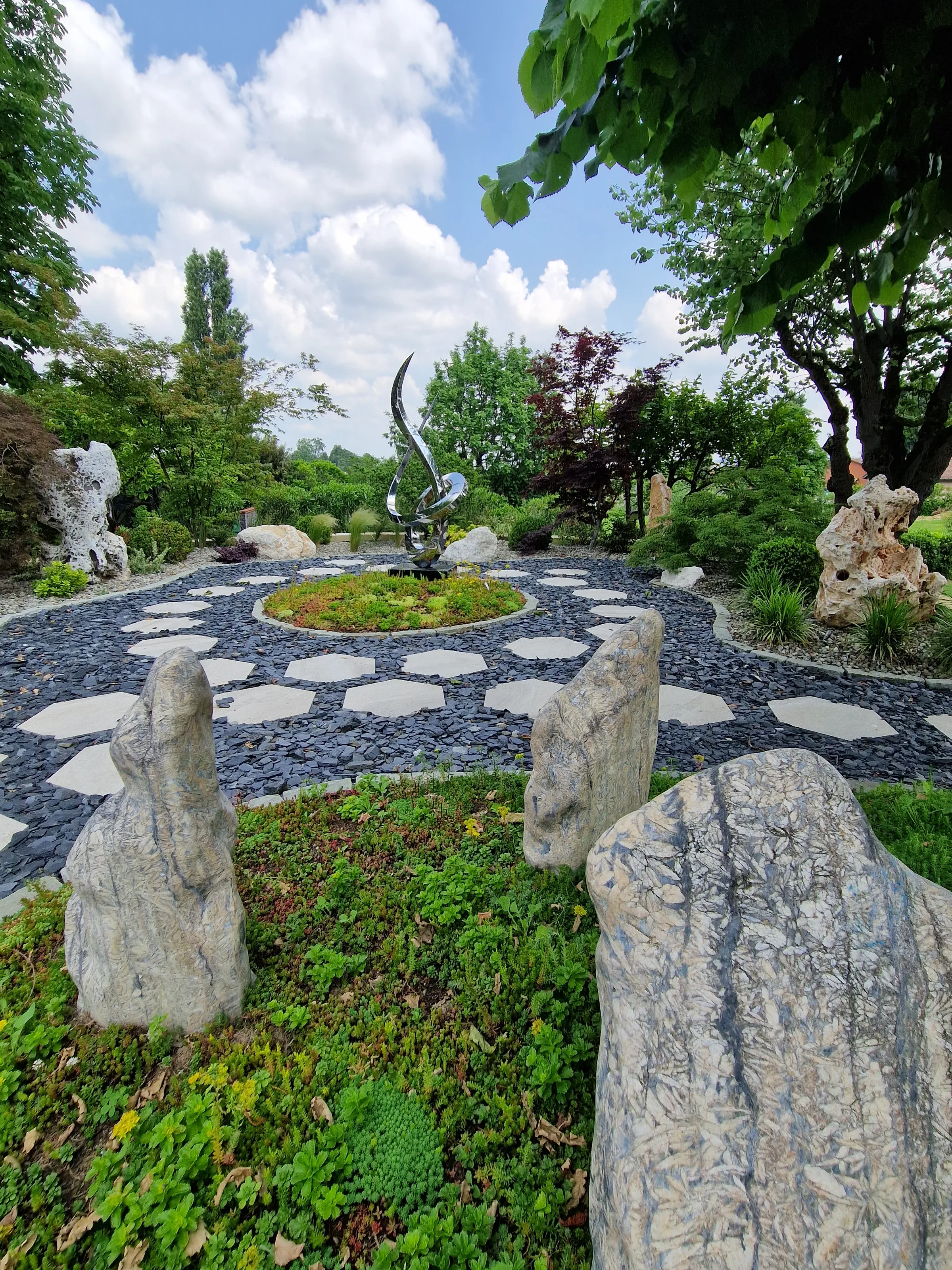 Passi esagonali in pietra chiara su un letto di scaglie d'ardesia, con menhir di pietra piantati nel tappezzante e una scultura in metallo al centro