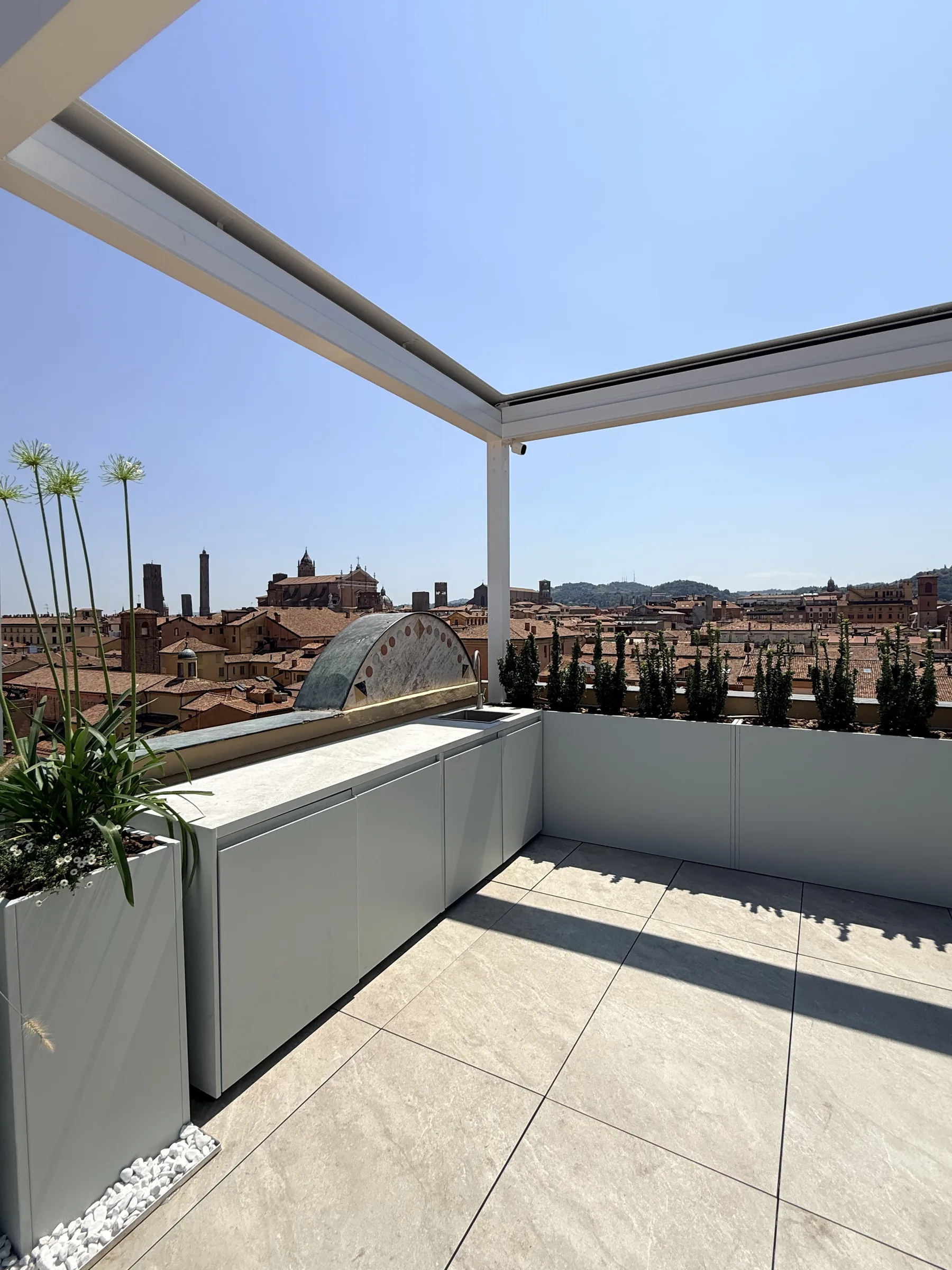 Terrazzo sui tetti di Bologna con la pergola bianca, la cucina da esterno, le fioriere grigie con gli arbusti colonnari e le torri sullo sfondo