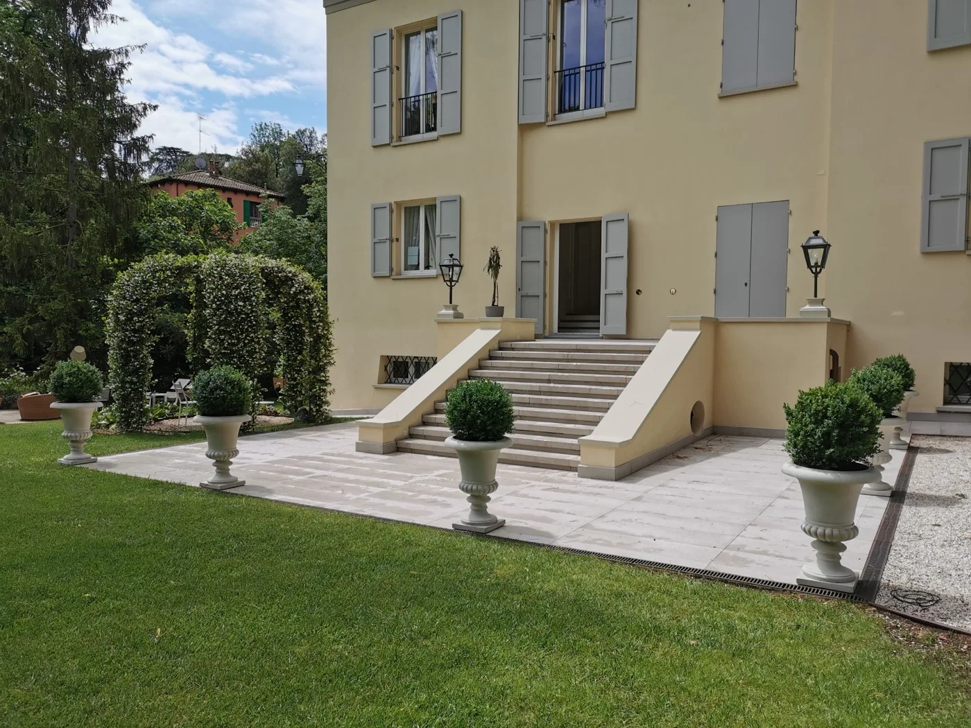 Fronte di una villa con scalinata in pietra, arco di gelsomino potato, vasi a calice con sfere di bosso e prato rasato attorno