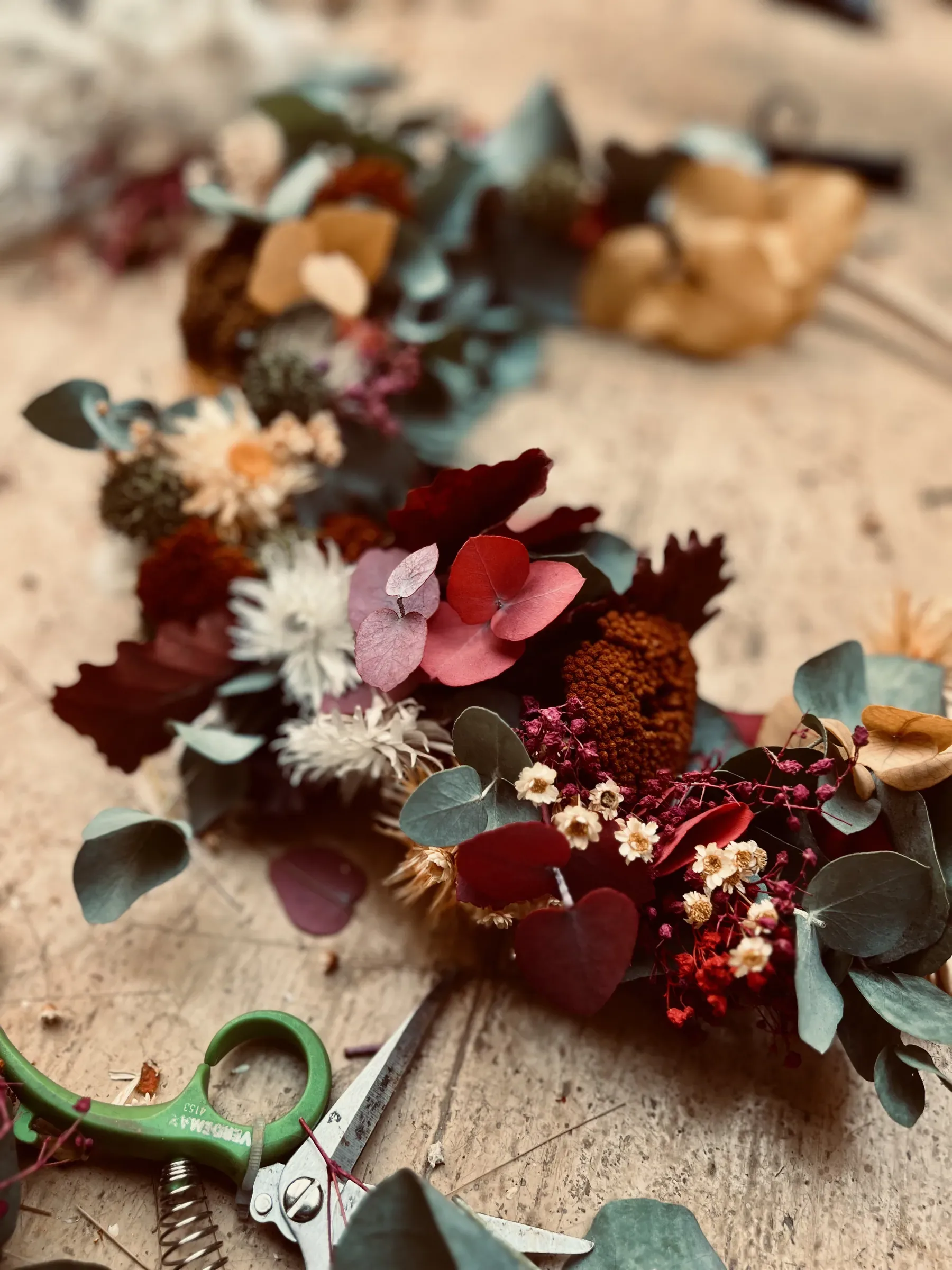 Una corona di fiori secchi in lavorazione sul tavolo di legno: eucalipto, foglie rosse, margheritine color crema e rametti di fiorellini rossi, con le forbici accanto