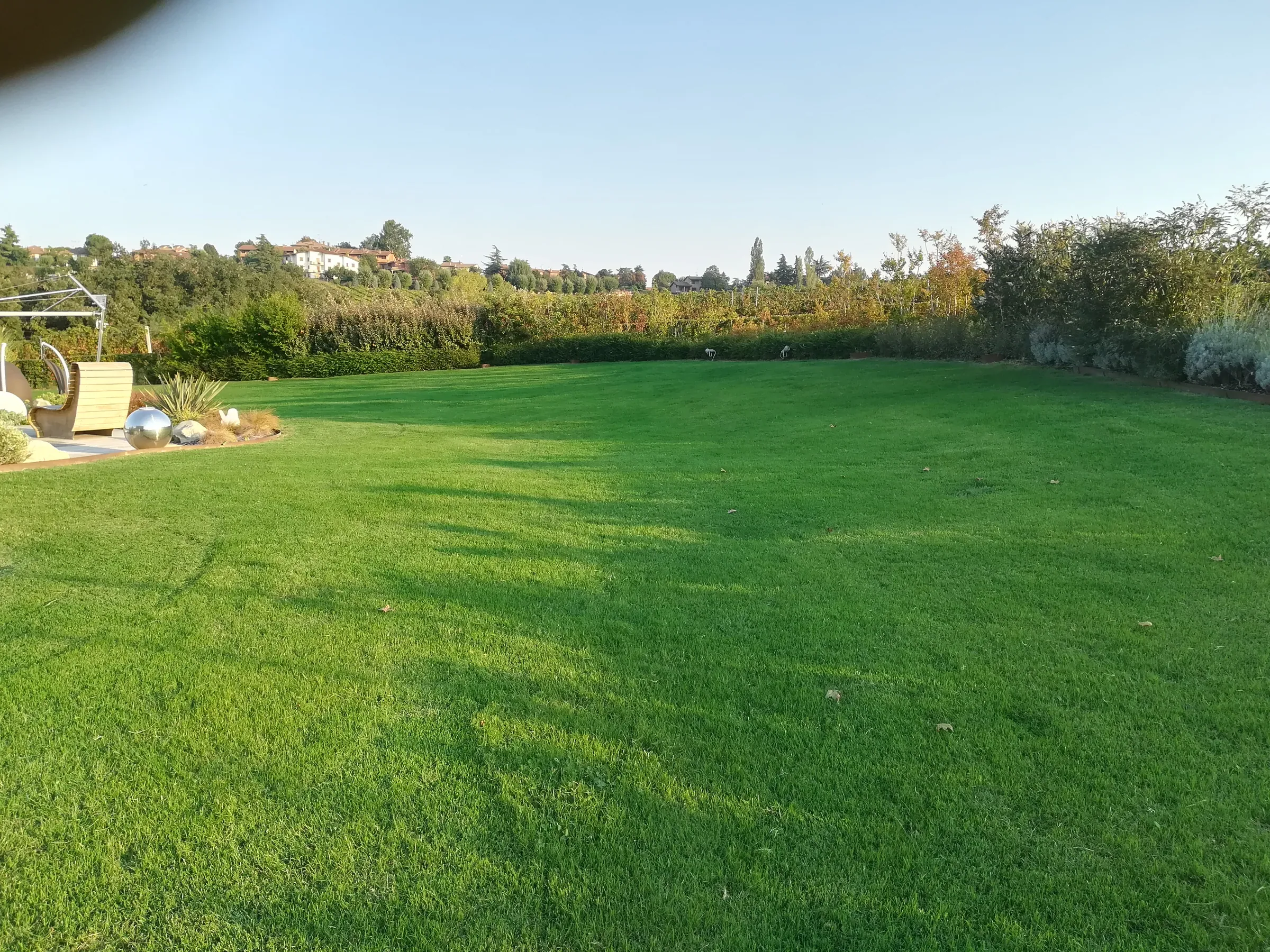 Grande prato rasato nella luce radente, chiuso da una siepe mista, con una seduta e sfere in acciaio in un angolo e le colline sul fondo