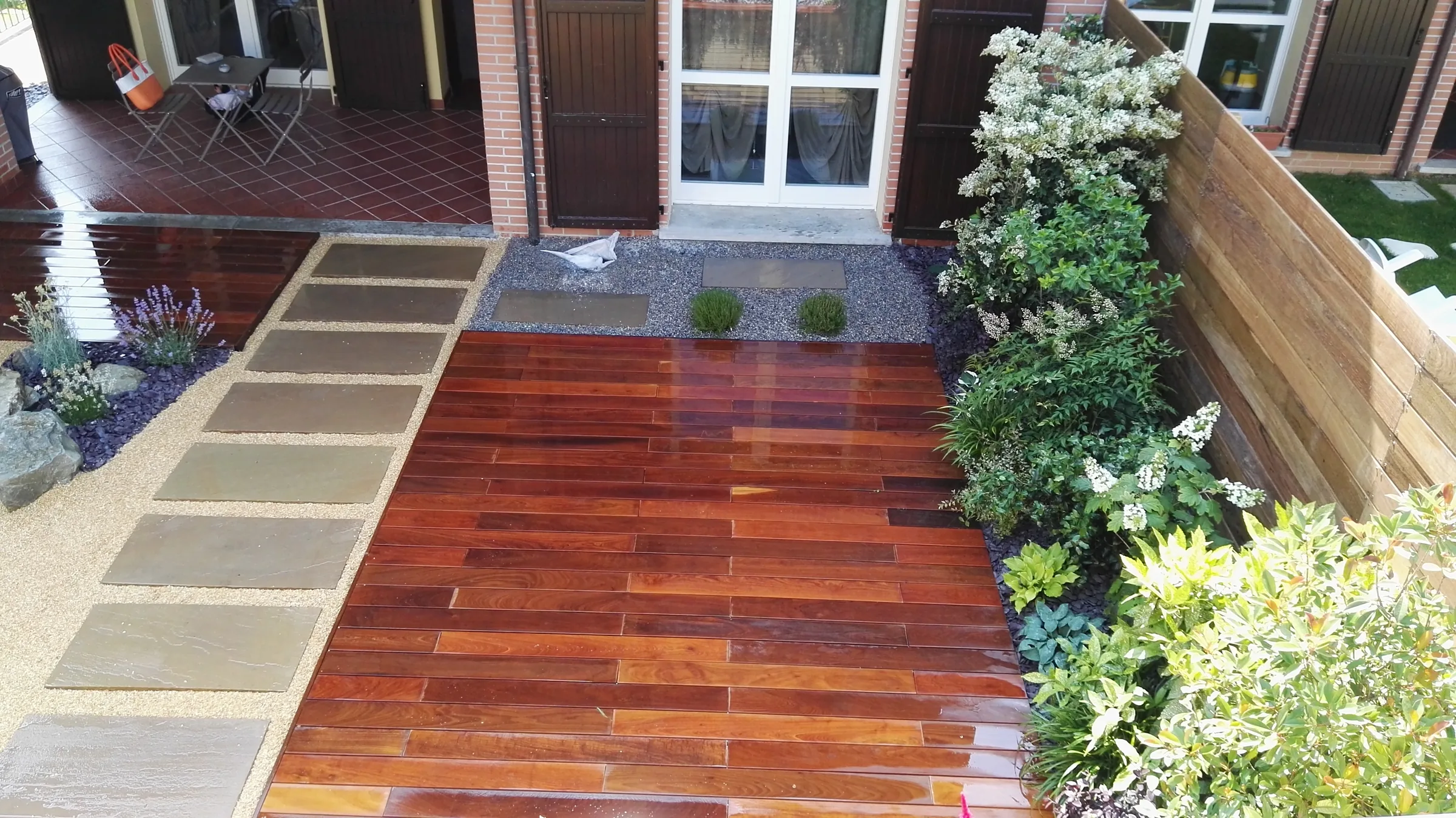 Pedana in listoni di legno rossiccio lucido, con un camminamento di lastre grigie nel ghiaietto a lato e aiuole di arbusti lungo una recinzione in legno
