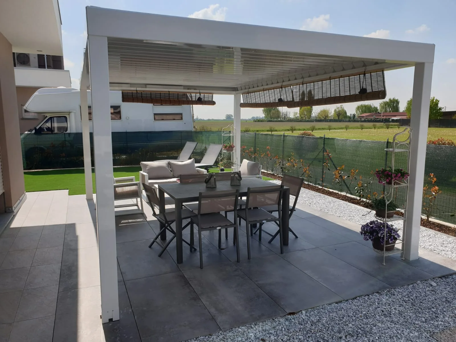 Pergola bianca con tavolo e sedie su un pavimento scuro, fra il prato e un campo aperto dietro la recinzione