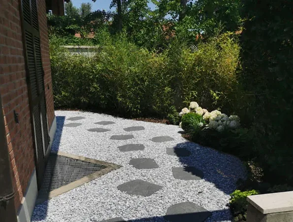 Dopo: passi in pietra scura su ghiaia bianca accanto al muro in mattoni della casa, con ortensie bianche e una siepe alta sul fondo