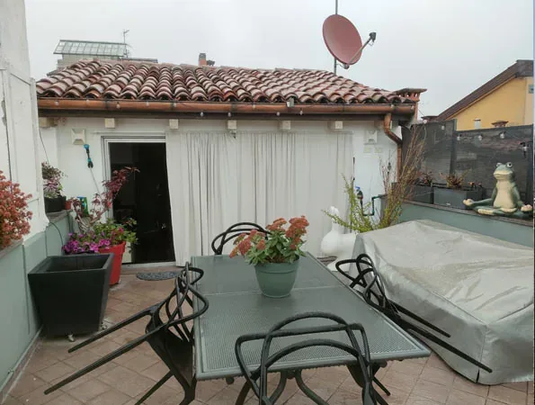Prima: tavolo di metallo con un vaso di fiori arancio, un mobile coperto da un telo, vasi fioriti e una tenda bianca accanto alla porta