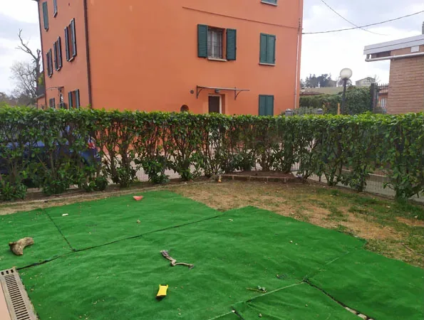 Prima: pezzi di erba sintetica accostati sul terreno lungo la siepe, con la casa arancione sul fondo