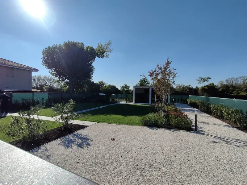 Giardino con vialetti in ghiaia, prato, aiuole di arbusti e una pergola bianca con vetrate sul fondo